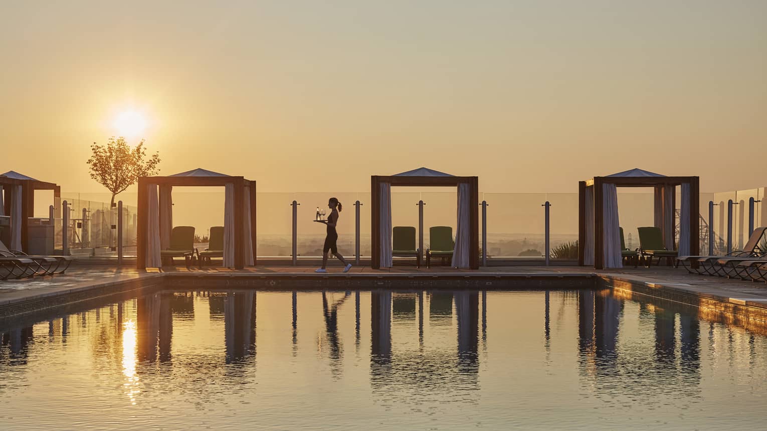 Silhouette of woman walking with tray with wine carafe and glasses, by rooftop pool at sunset