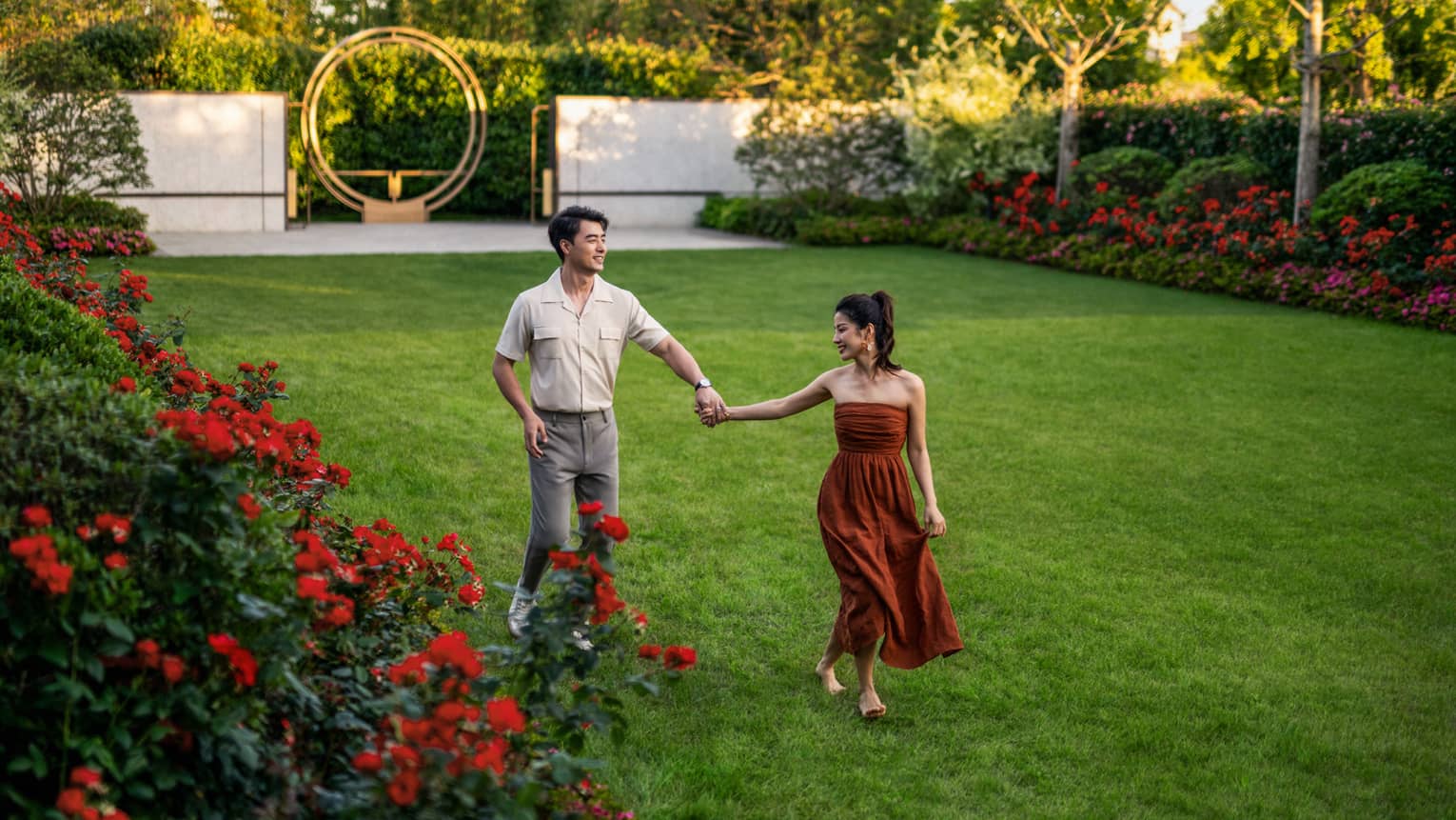 A couple holding hands crosses a thick lawn bordered by trees and bright red and fucsia blooms, and a metal abstract artwork.