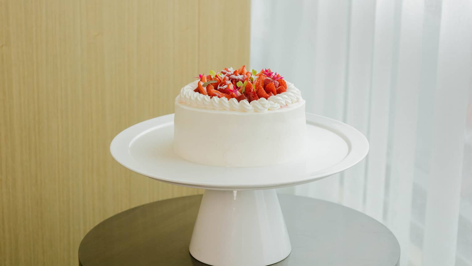 Round strawberry shortcake with white icing and topped with fresh sliced strawberries is set atop a round white cake stand