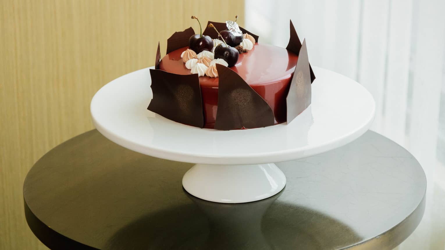 Round black forest cake with a red glazed center, jagged pieces of chocolate lining the sides, and fresh cherries and dollops of mousse is set atop a round white cake stand