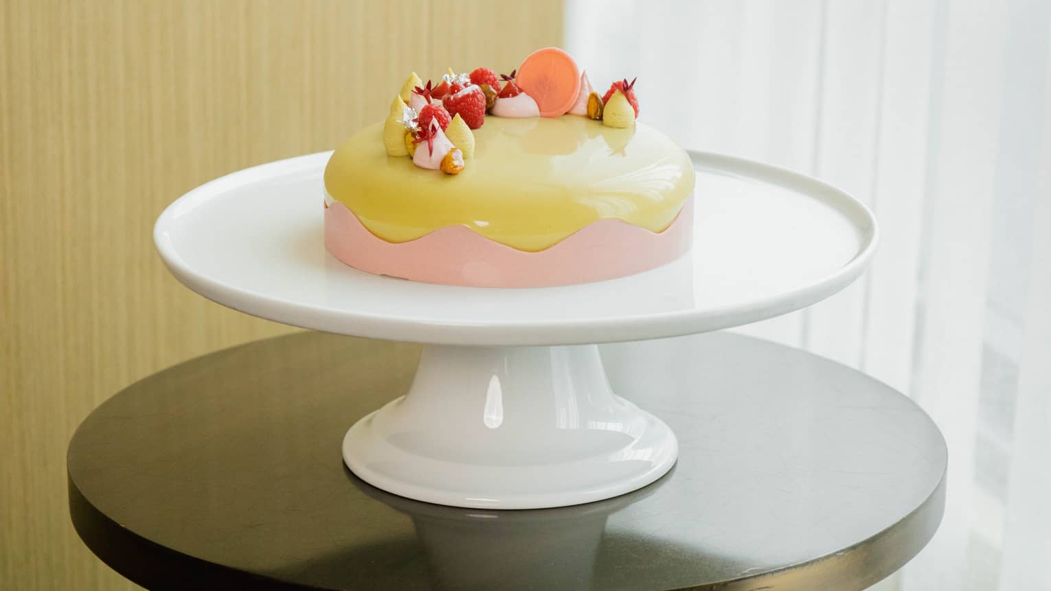 Round cake with a glazed pistachio-colored icing, a pink scalloped-edge border around the side and a candied-fruit topping is set atop a round white cake stand