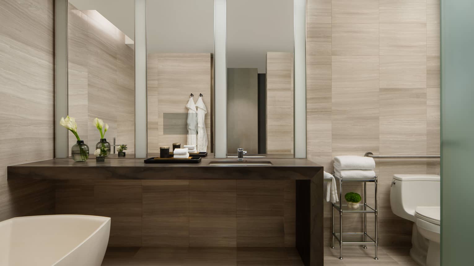Luxury hotel bathroom with dark wood vanity, wide mirror, freestanding tub, glass vases with flowers, toilet and stacked towels on a metal rack