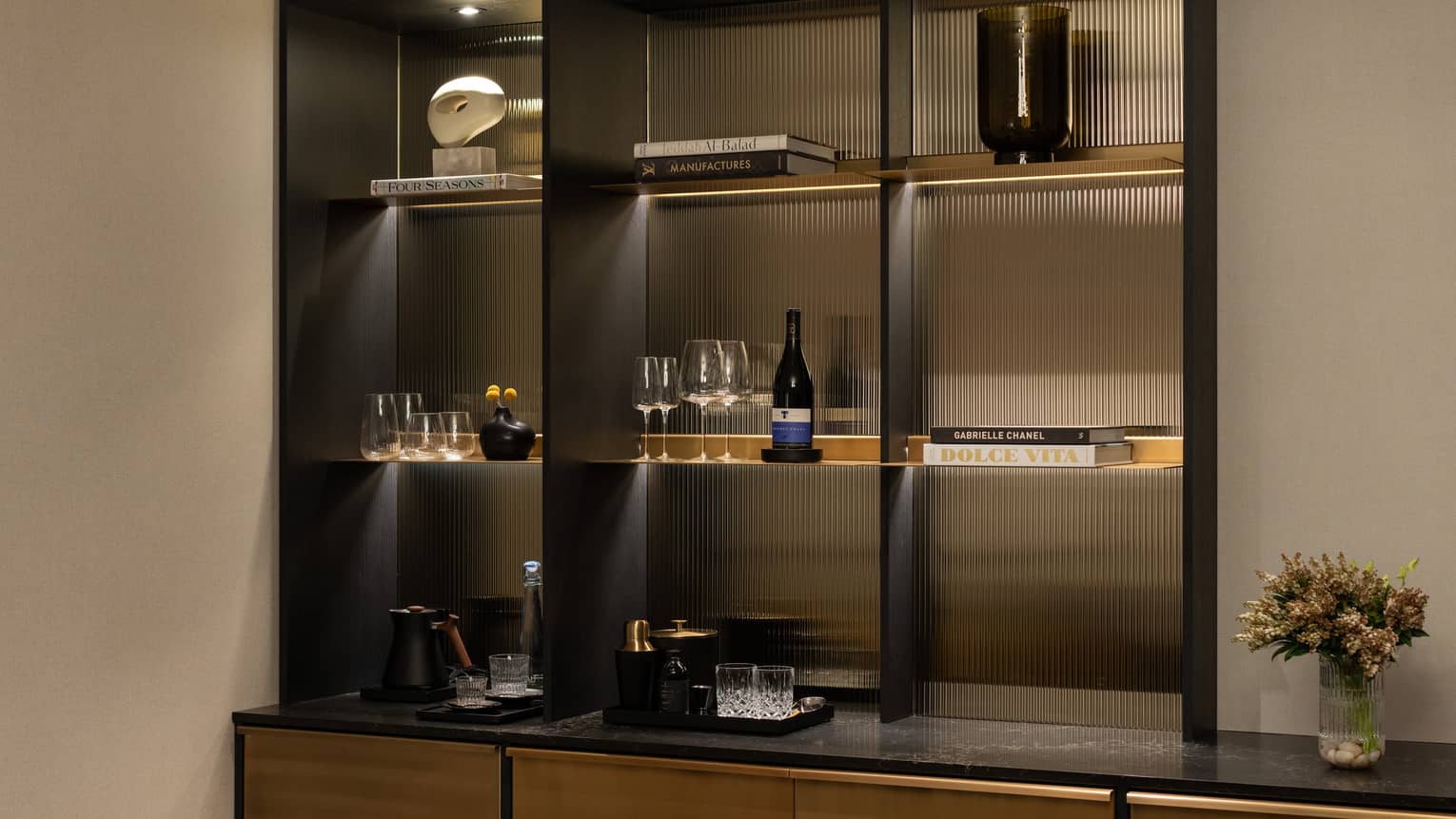 Hotel suite bar shelf with books, glasses, wine bottle, decorative pieces and a flower vase