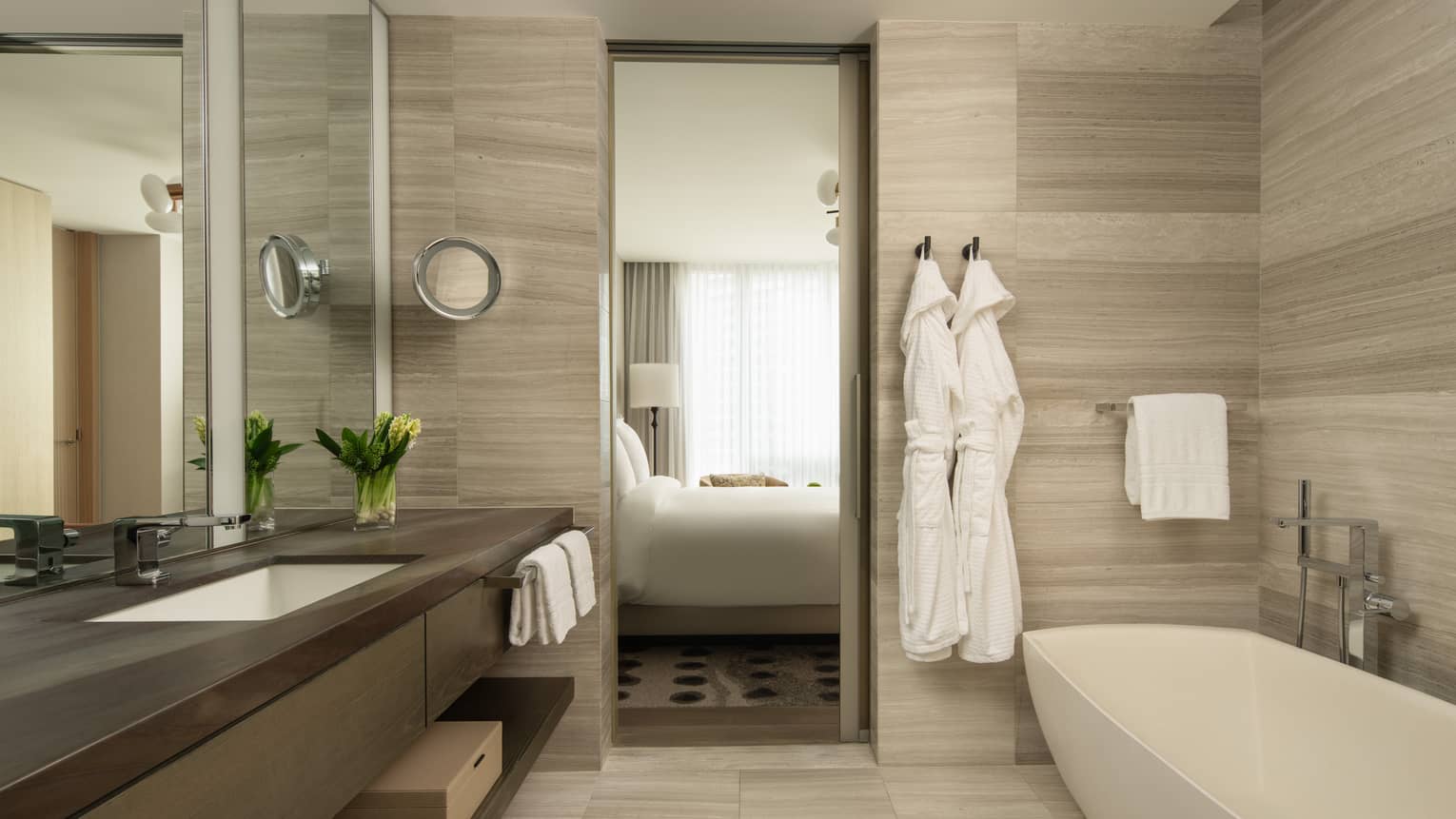 Luxury bathroom with long vanity, a freestanding bathtub, wood accents, and two bathrobes hanging, overlooking the bedroom through a sliding door 