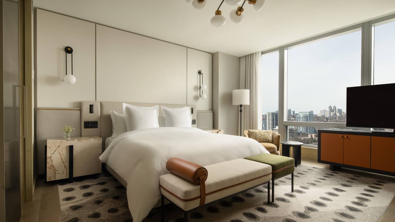 Hotel suite bedroom with a king bed, nightstands, patterned carpet, bench, armchair, large windows and a TV