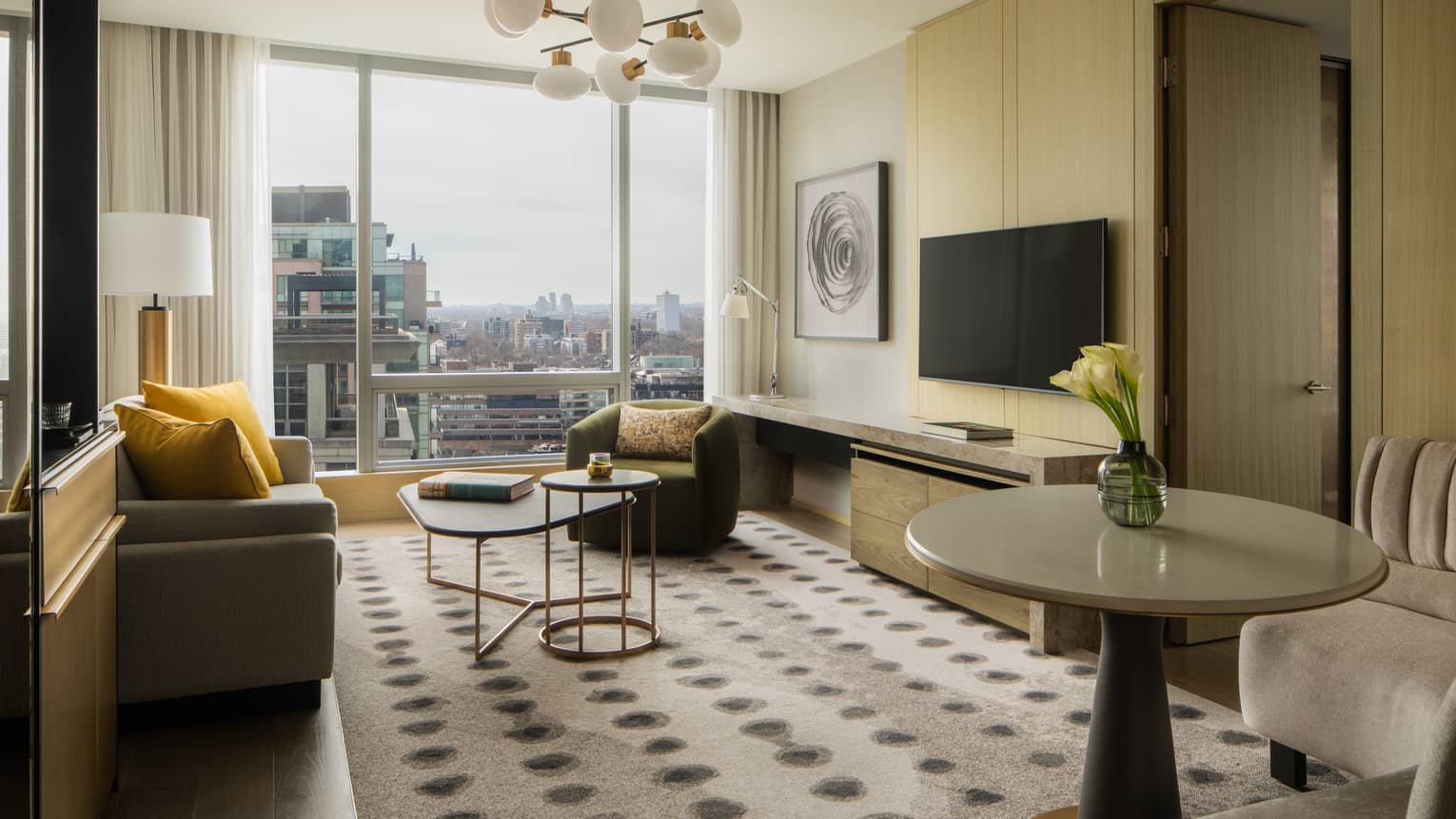 Hotel suite living room with a beige sofa, yellow pillows, armchairs, coffee tables, wall-mounted TV, console table, abstract artwork and large windows 