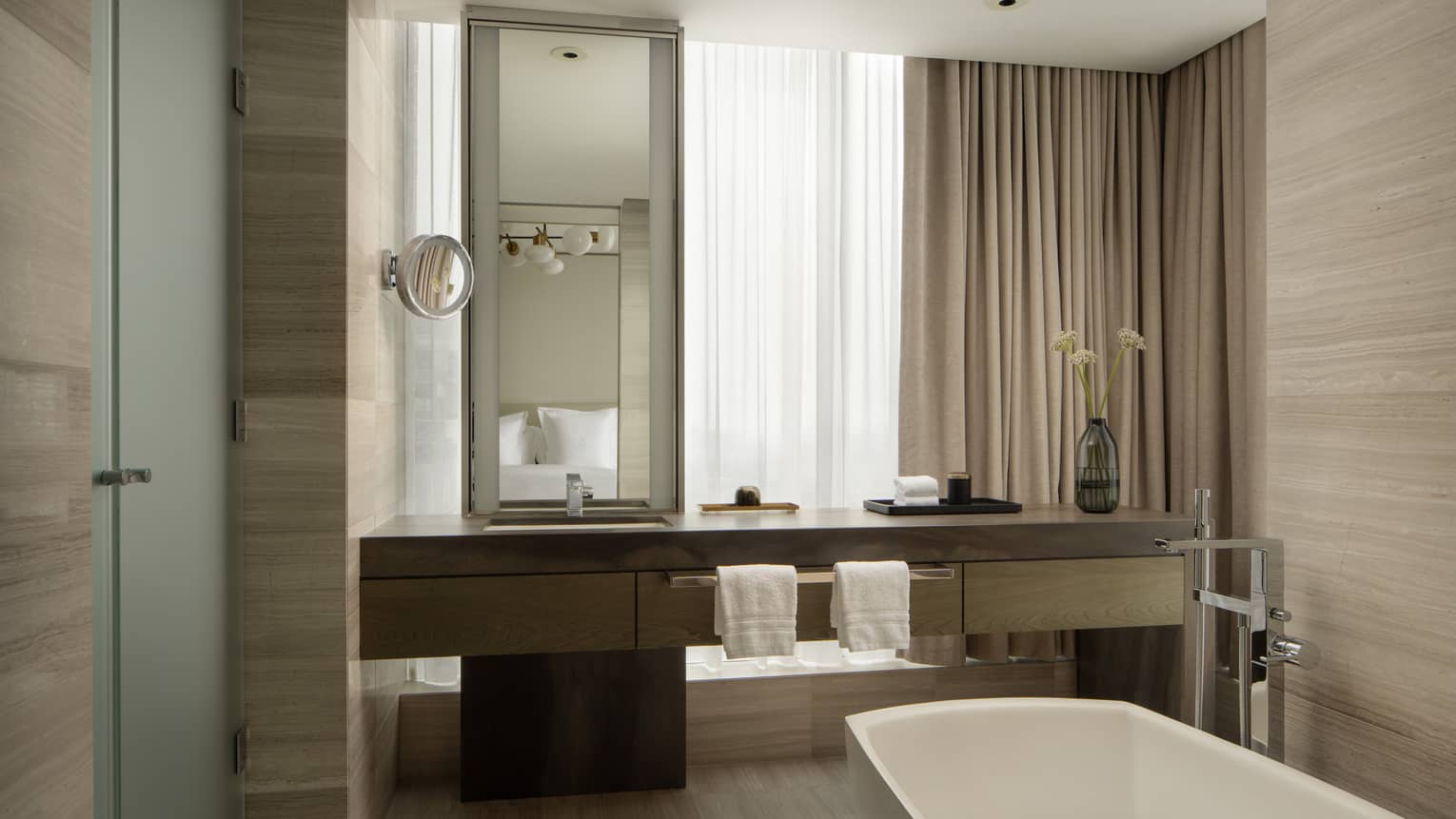 Hotel suite bathroom with dark wood vanity, large mirror, walk-in shower, freestanding bathtub, two towels and tall beige curtains