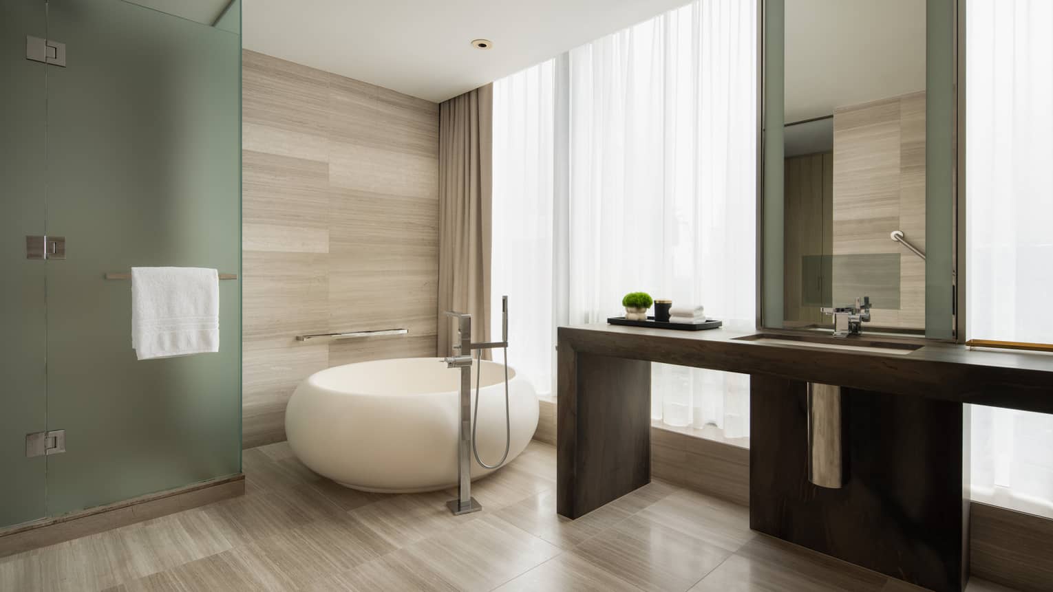 Luxury hotel bathroom with round freestanding tub, glass shower with towel bar, long wooden vanity, mirror and sheer window curtains
