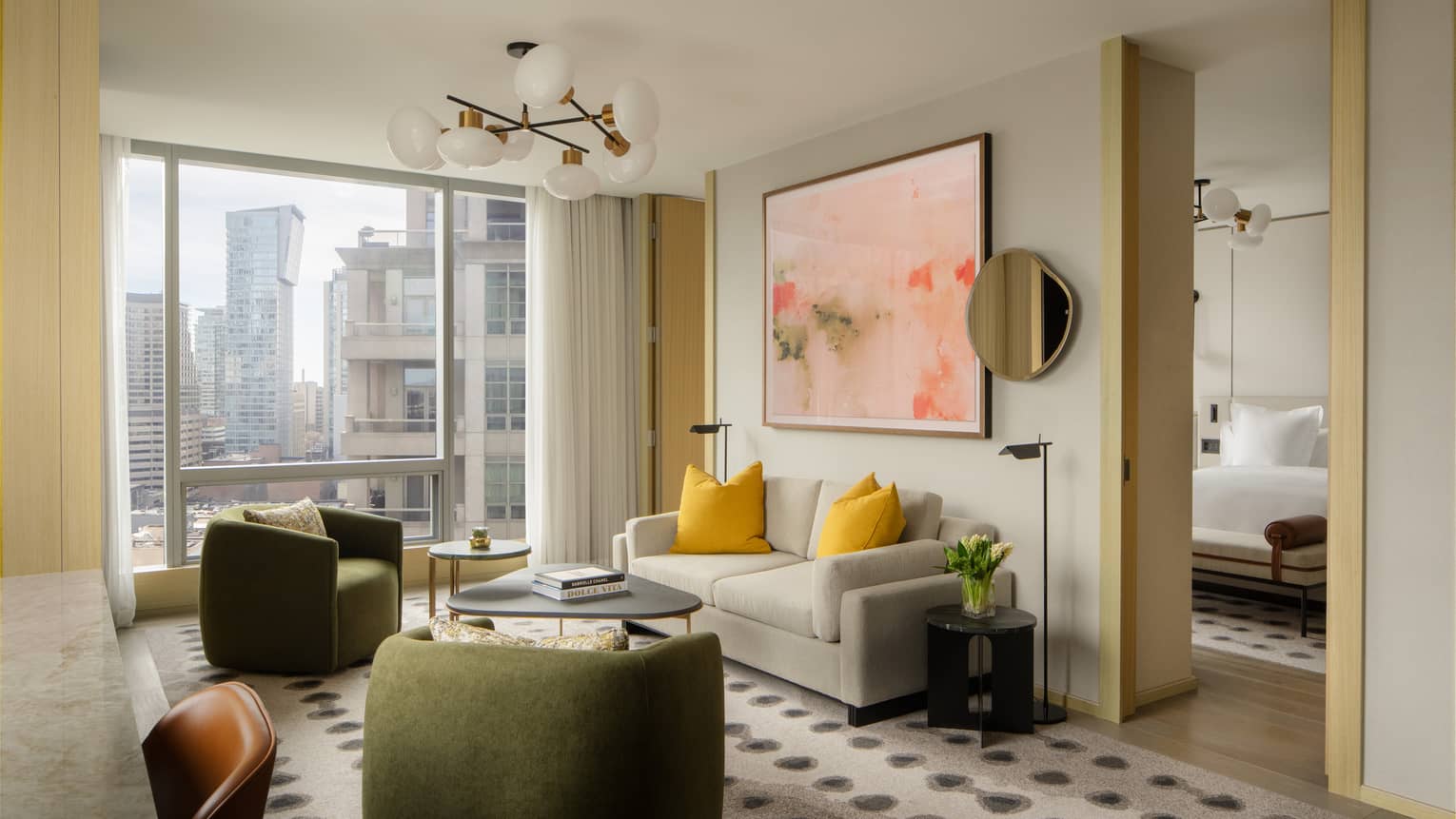 Living area with a beige sofa, yellow accent pillows, green armchairs, abstract artwork, and large windows with city views, connected to a bedroom