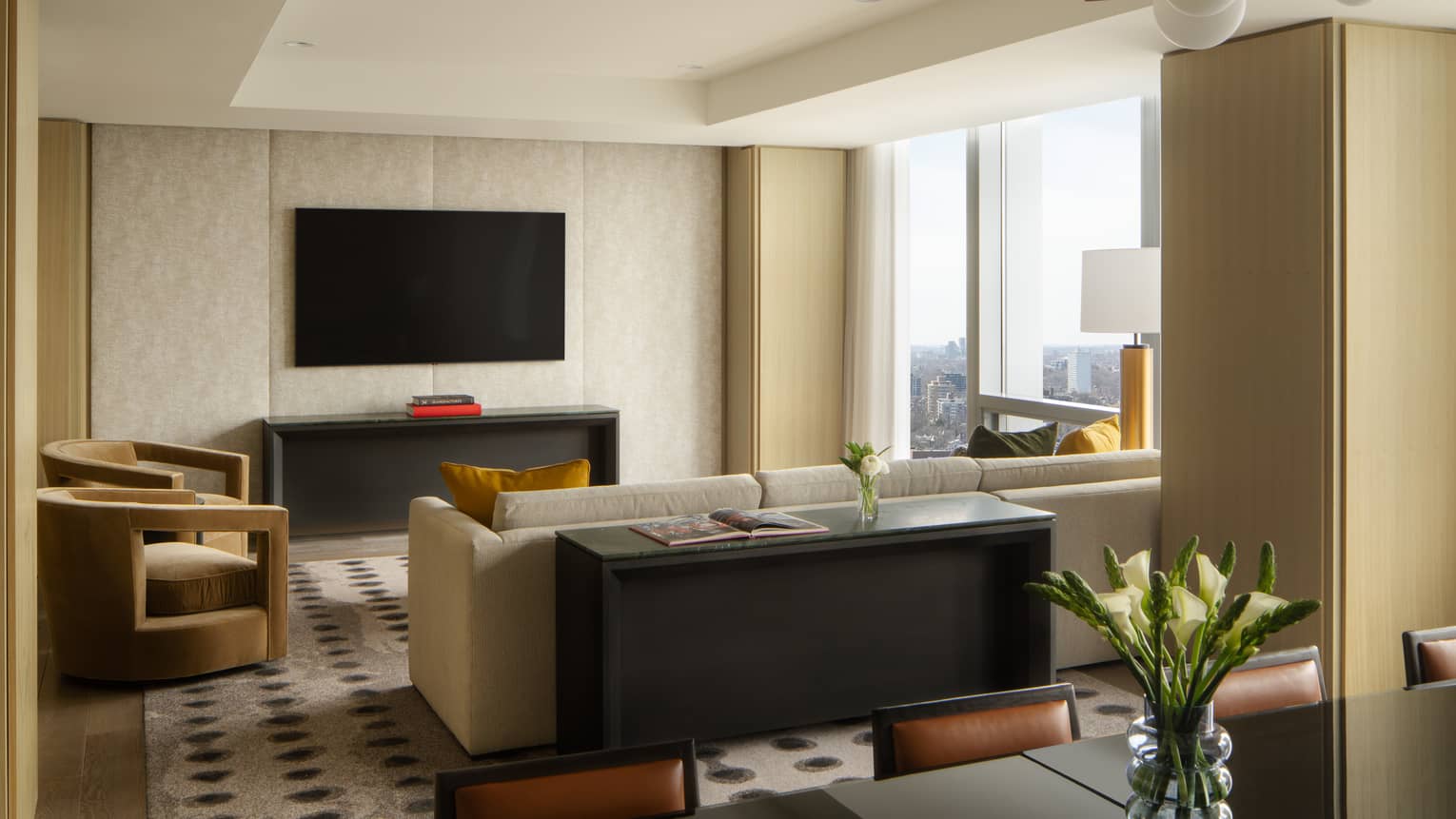 Luxury suite living area with a wall-mounted TV above a black console, beige sectional sofa, armchairs, flower vase and large windows 