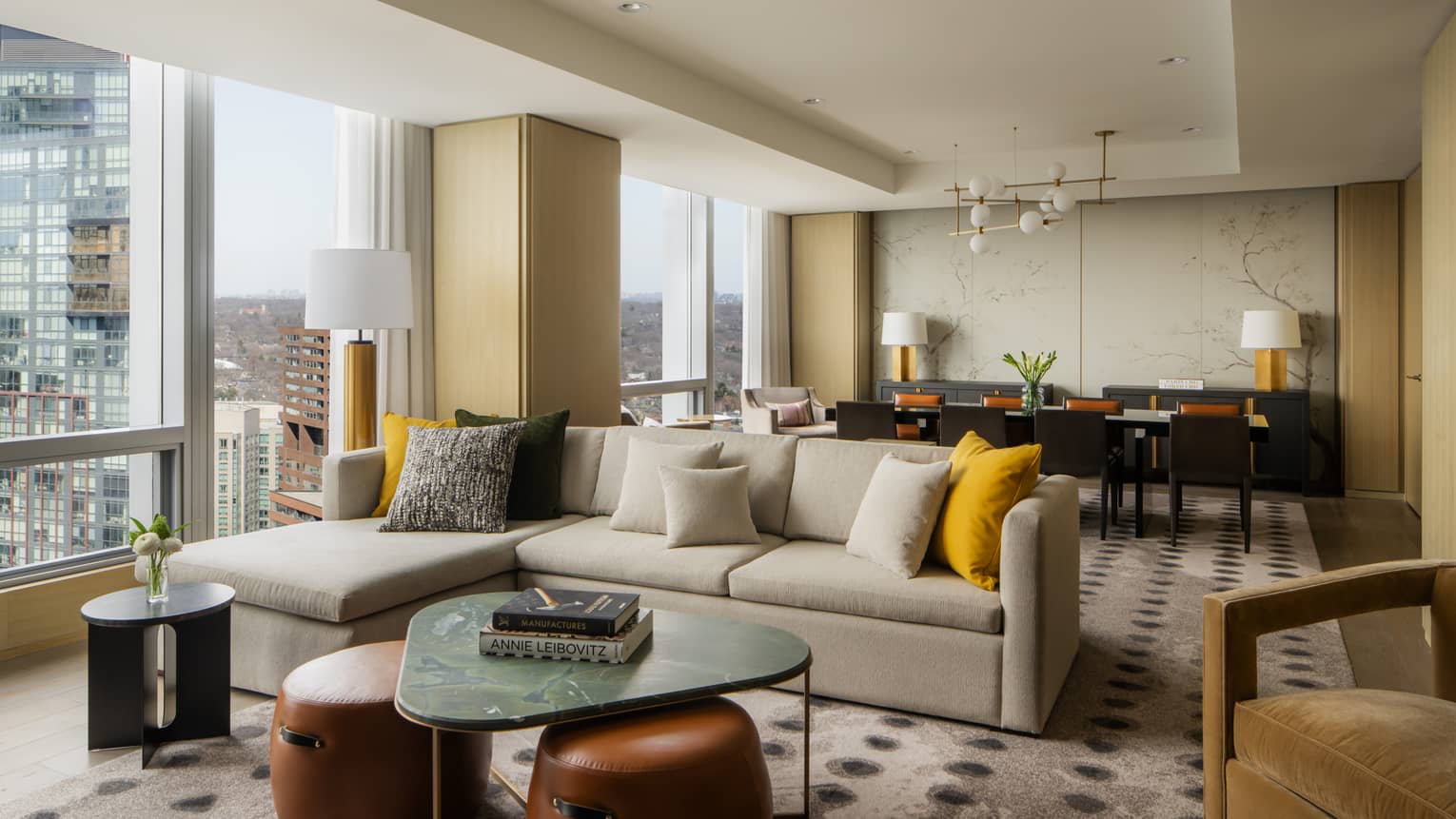 Spacious suite living area with a large sectional sofa, yellow and gray pillows, marble coffee table, and a dining area with elegant décor and city views