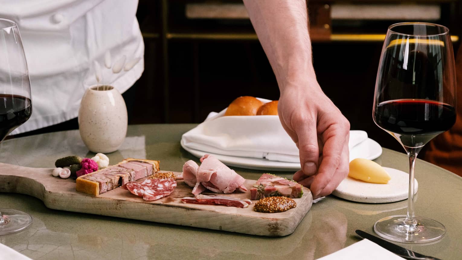Chef places a wooden board of charcuterie on table next to glass of red wine