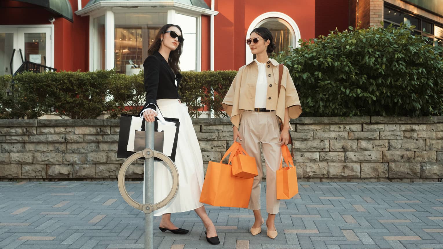 Two stylish people with shopping bags, standing outdoors in front of a red building, dressed in chic, neutral-coloured outfits and sunglasses