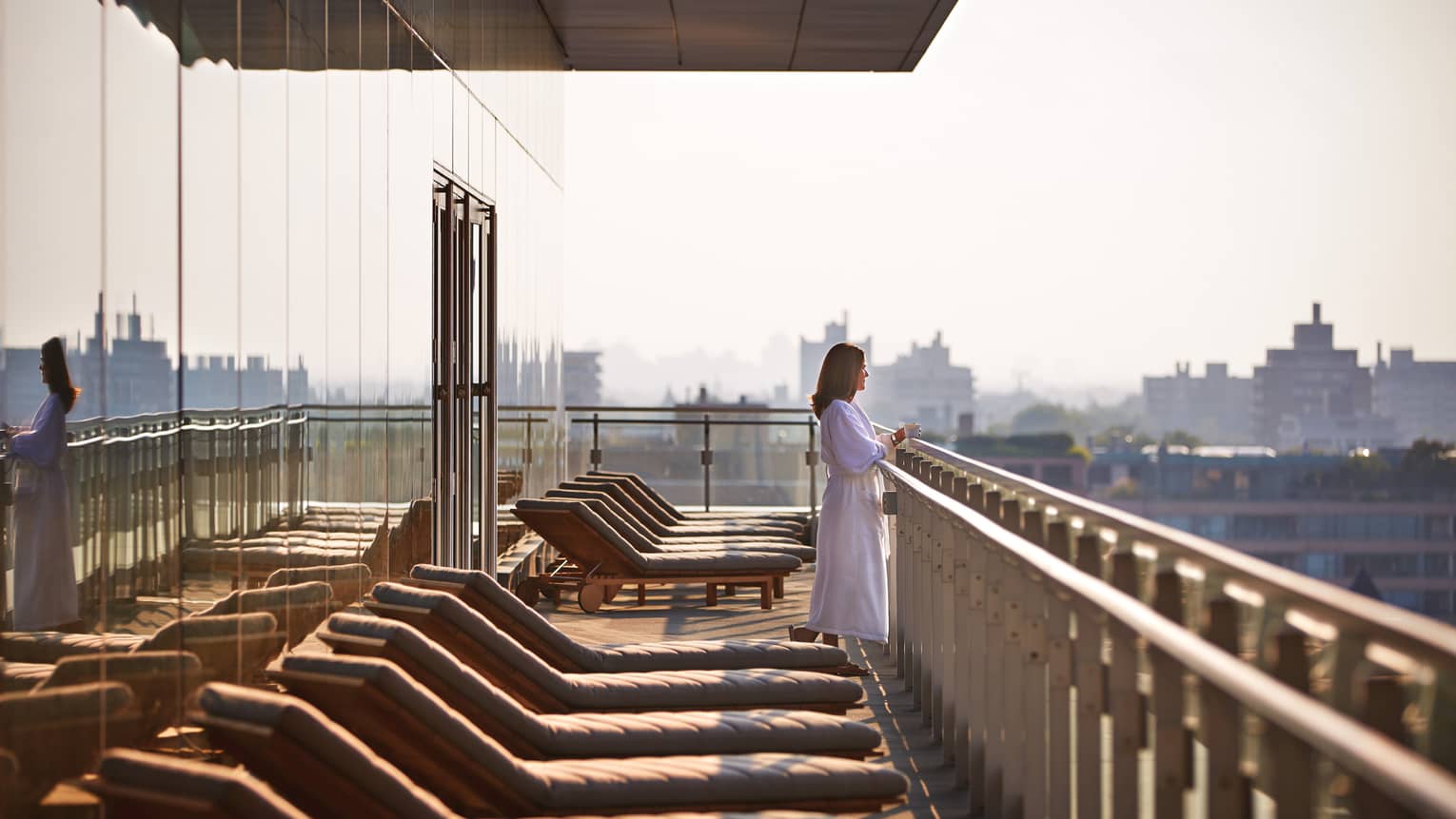 Woman in white bathrobe looks out over balcony ledge near row of spa patio chairs 