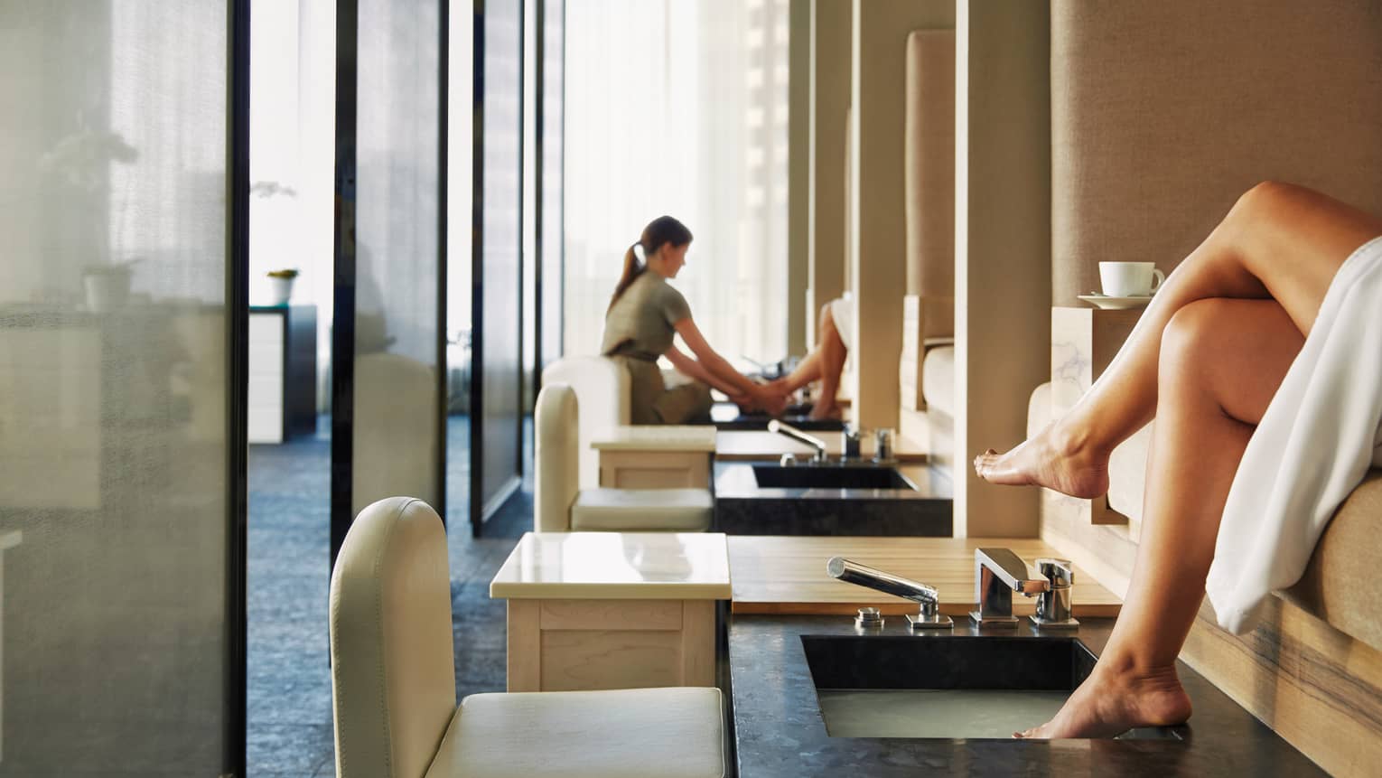 Woman's feet on pedicure sink along row of spa chairs, salon staff in background