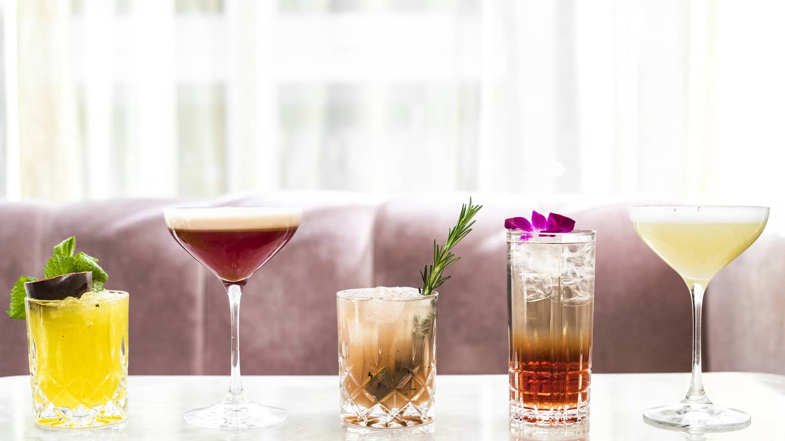 Five cocktails in crystal glassware on marble table before velvet chairs, sheer curtains 