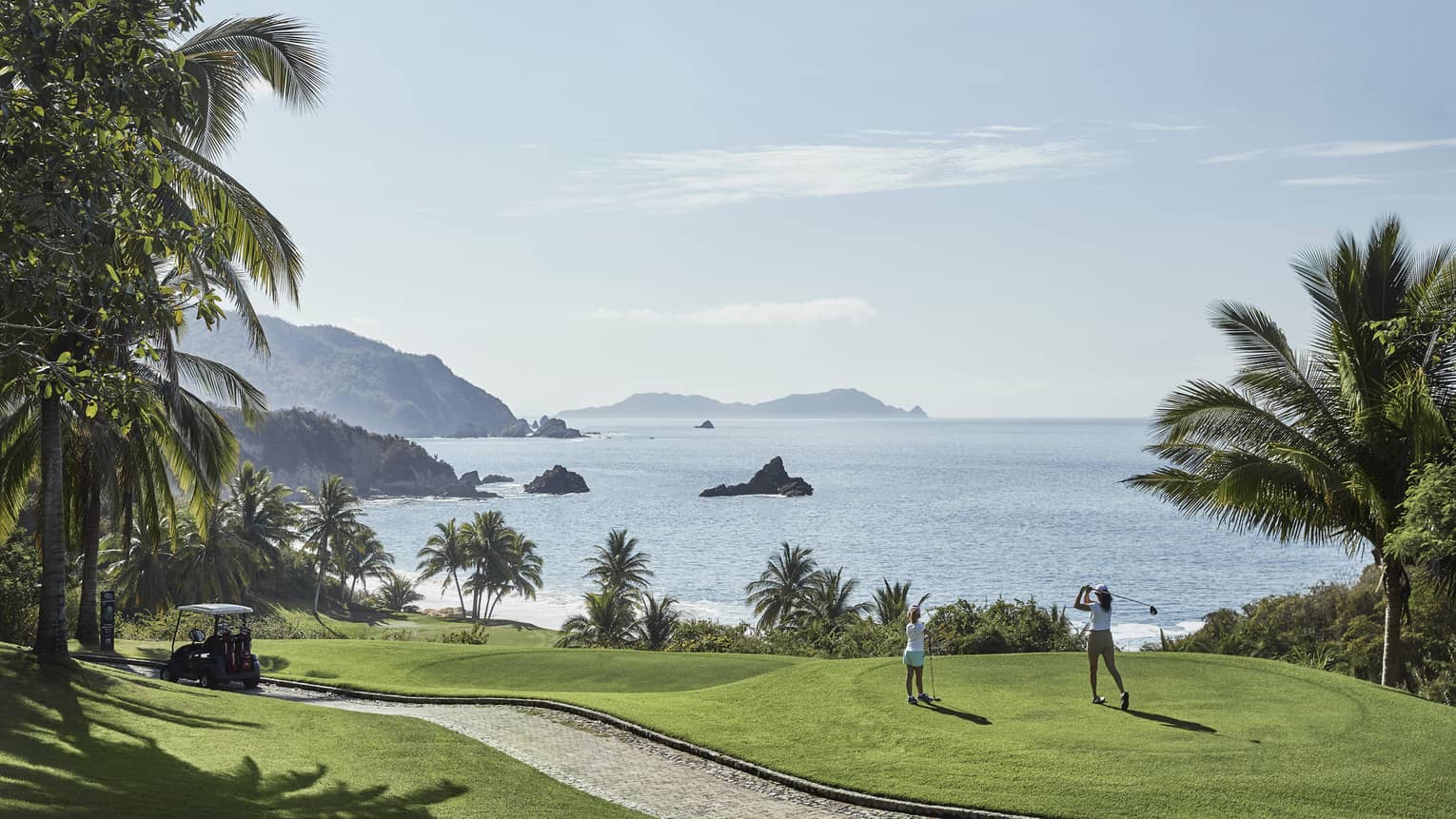 A guest swings from the tees on a hilly fairway bordered by palm trees and blue water with rocky outcrops as a child watches.