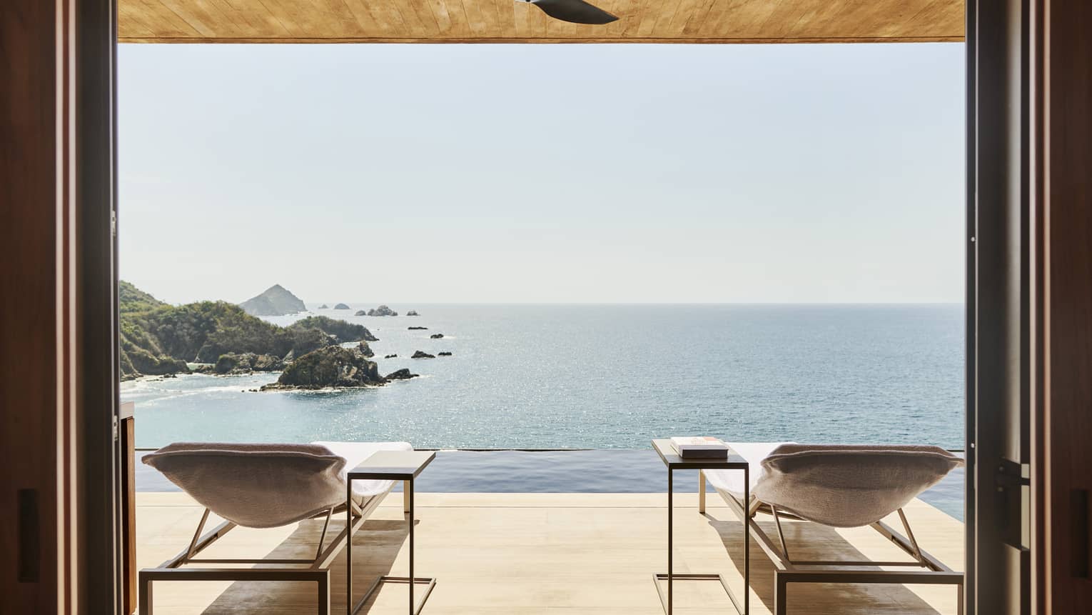 Open terrace with two lounge chairs and side tables, overlooking an infinity pool and a stunning ocean view with rocky islands. The space has a natural wood ceiling and serene ambience.