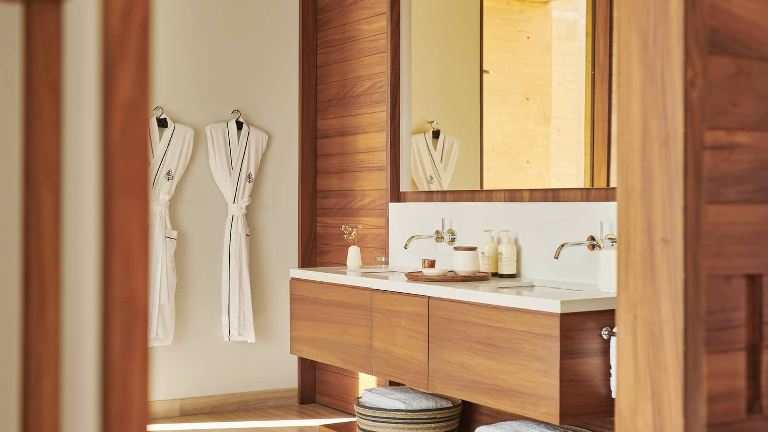 Modern bathroom with wooden accents, featuring a double-sink vanity, large mirror and neatly hung bathrobes. Baskets with towels are stored below the counter, creating a spa-like ambience.