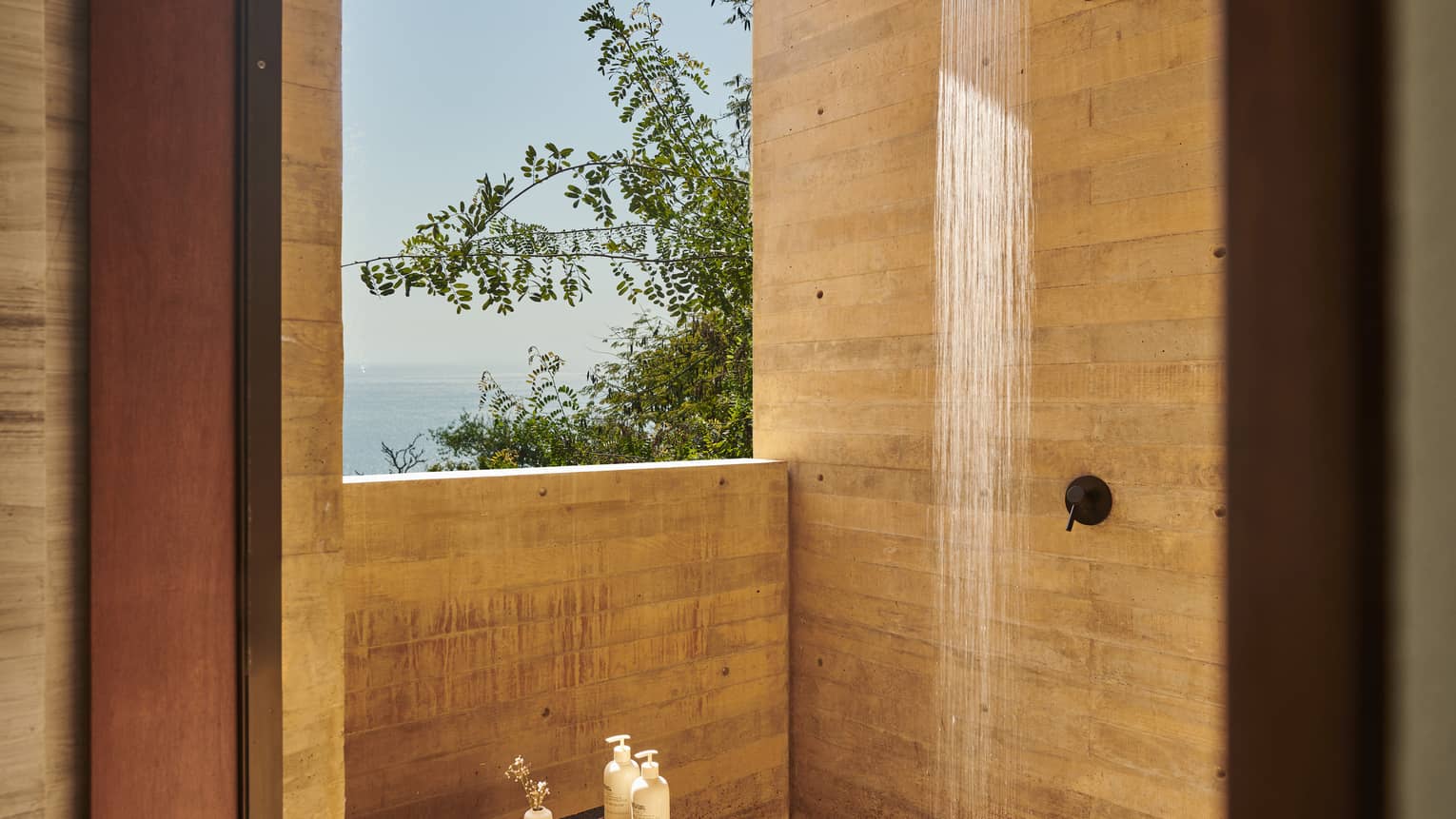 Open-air shower with natural wood walls, offering a view of lush greenery and the ocean. The sunlight enhances the serene, spa-like atmosphere.