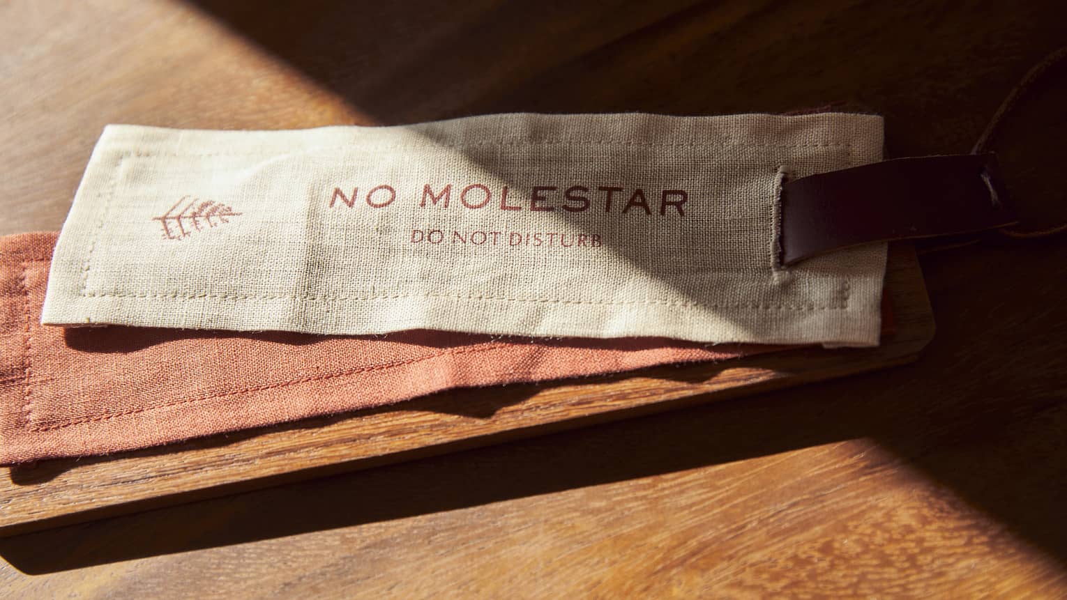 A fabric Do Not Disturb sign in Spanish (No Molestar) resting on a wooden surface with soft sunlight casting shadows across it