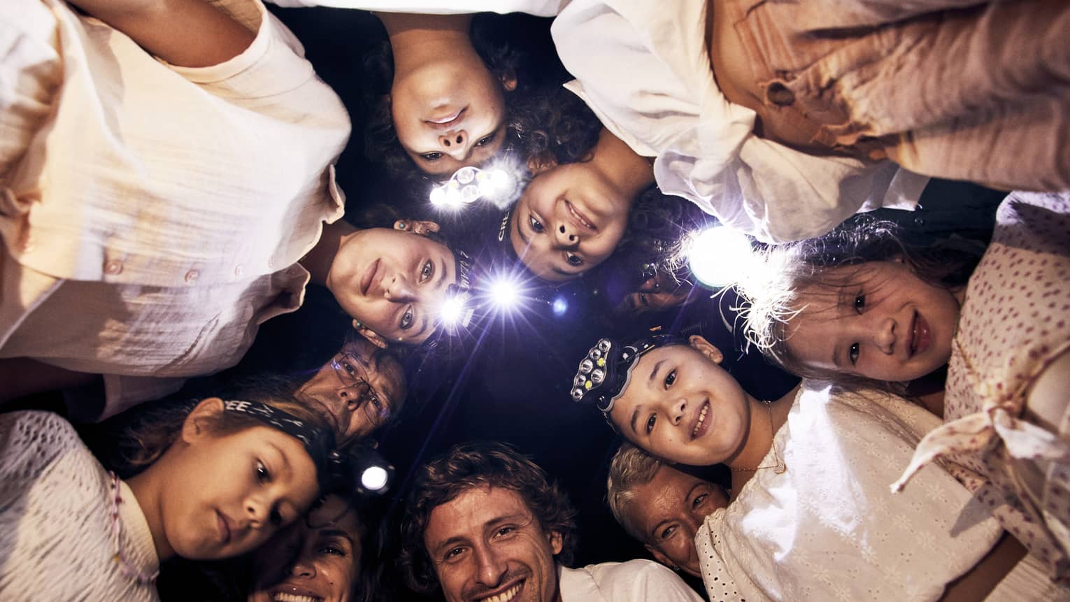A group of children and adults, some wearing headlamps, stand in a circle and smile downwards into the camera. 