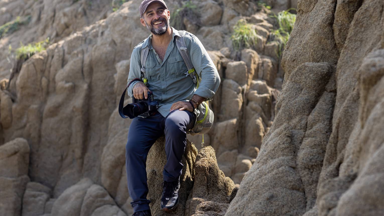 A photographer wearing a backpack and holding a camera in one hand sits atop a rock formation, smiling into the distance.