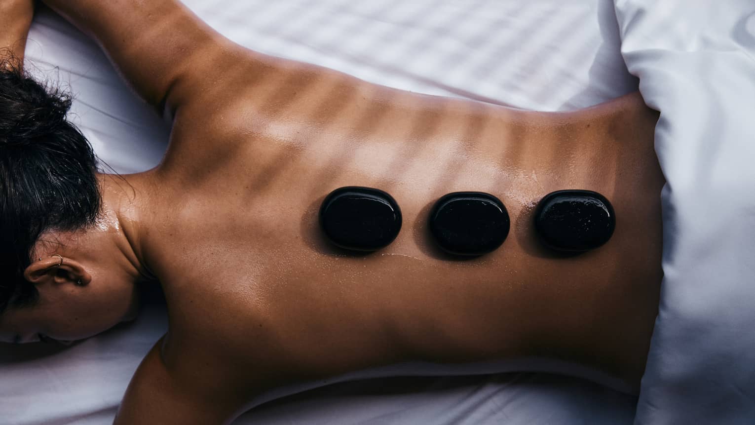 Close-up of a guest relaxing on their stomach on a bed of white linen with three black warming stones on their bare back.