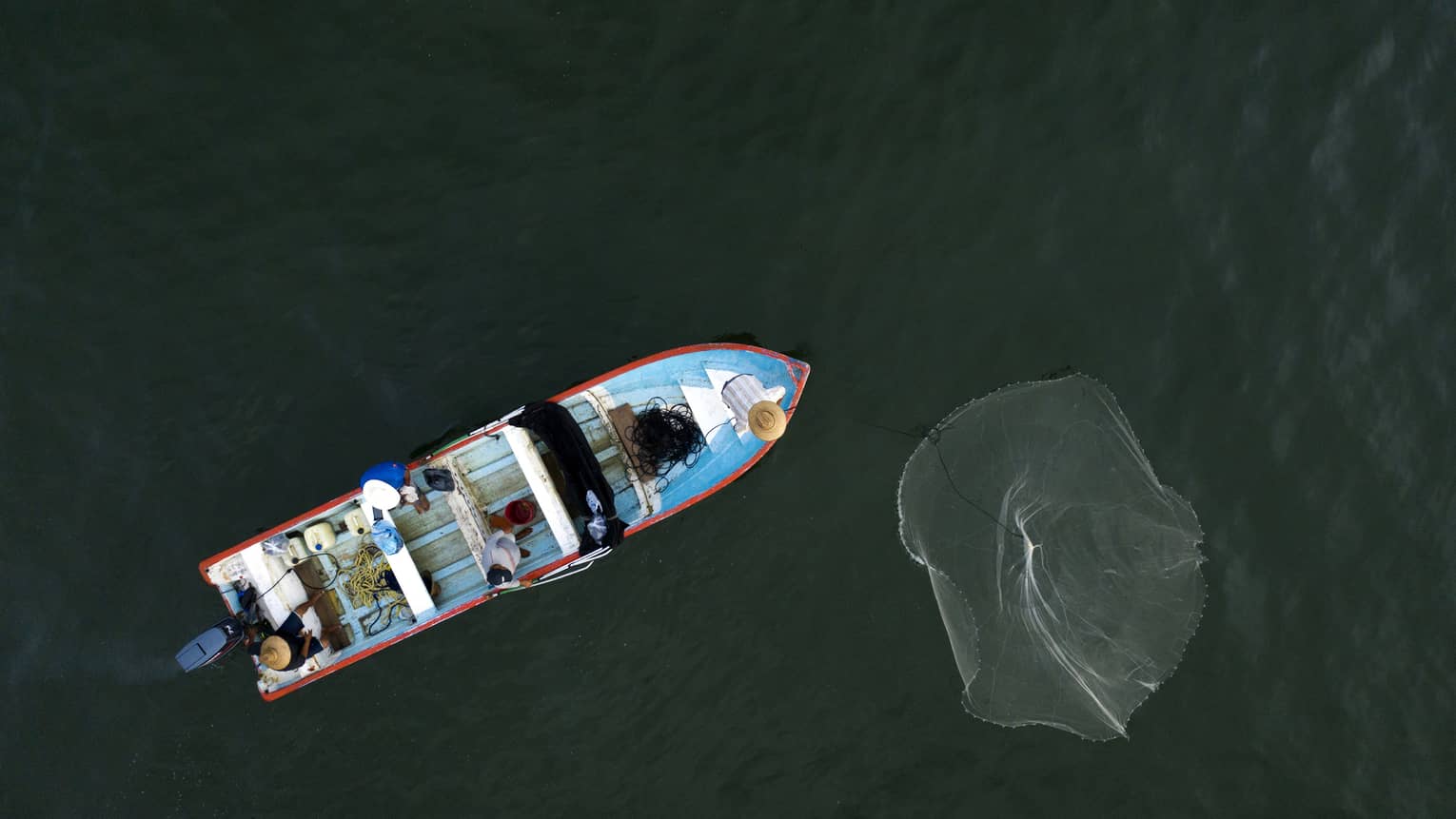 Overhead-view of a fishing boat on inky waters, a wide net cast off the bow, while three people aboard wait patiently. 