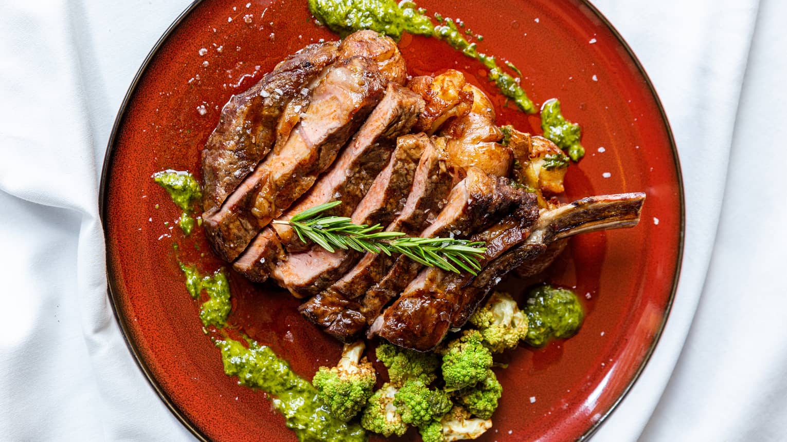 A red plate with sliced steak, herbs and a green sauce surround the steak.