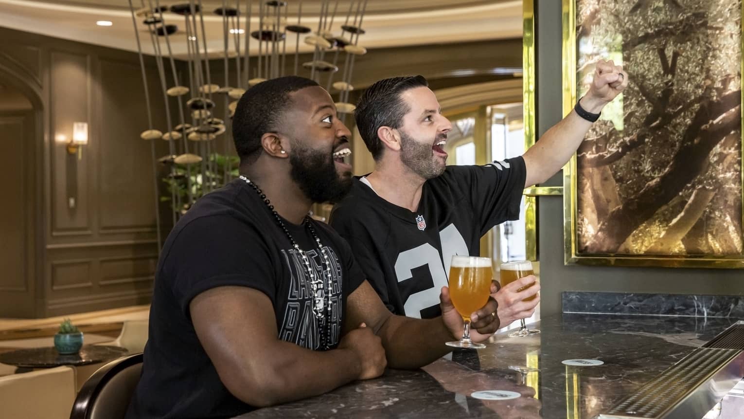 Two men in football shirts looking to the side while sitting at a bar with beer glasses in hand.