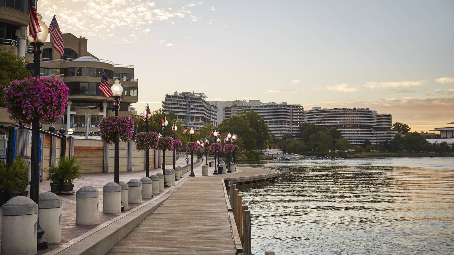 A waterfront with a wooden walkway next to light posts and flowers with building on the other side of a small road.