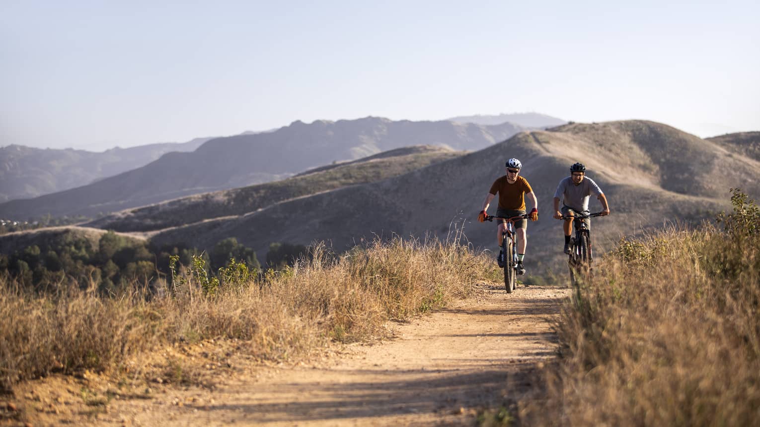 Two men riding bikes on a sand path between dry brush with hills behind them.