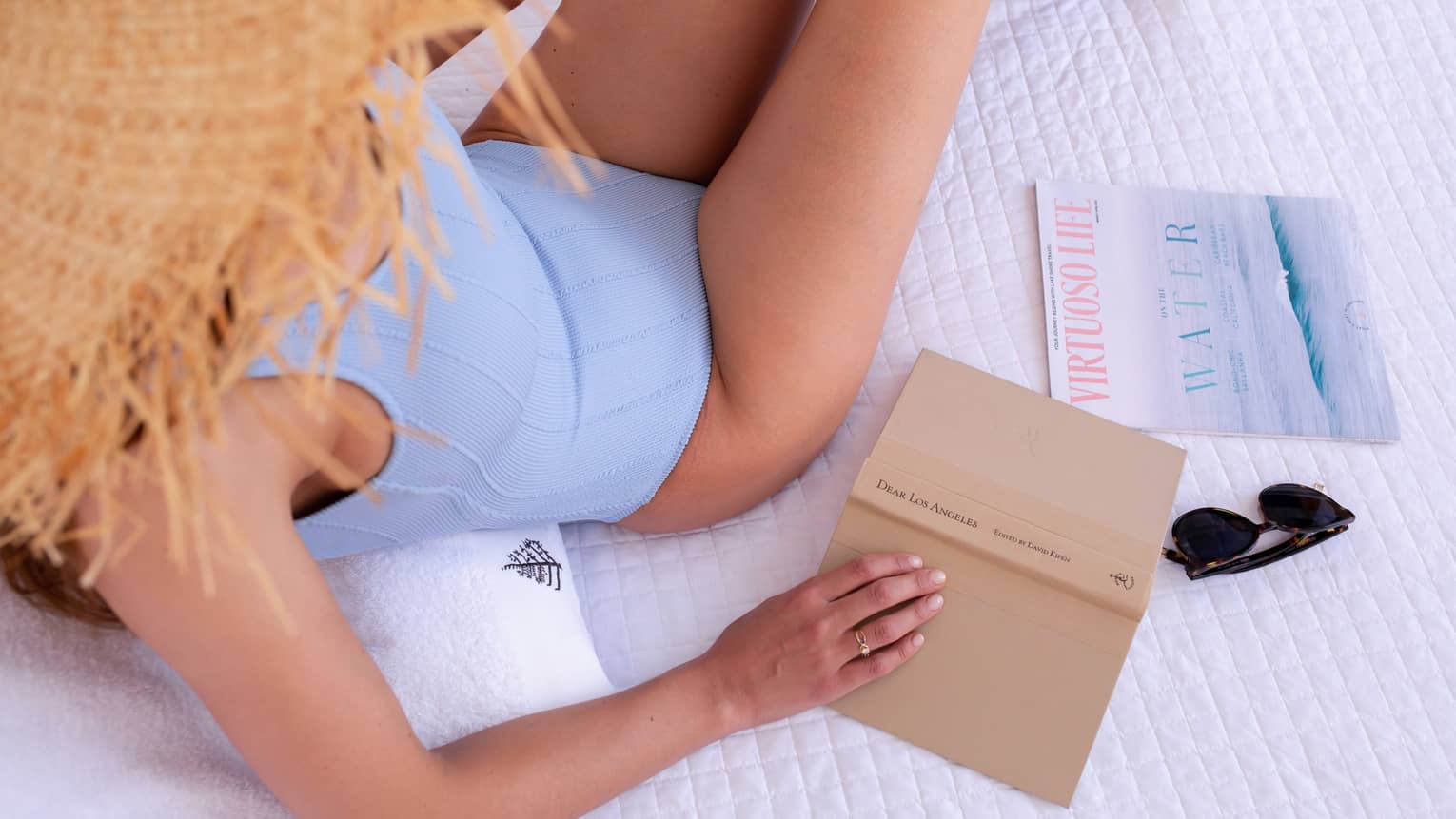 A woman in a blue swimsuit and straw hat holding a tan book while lounging on white bed