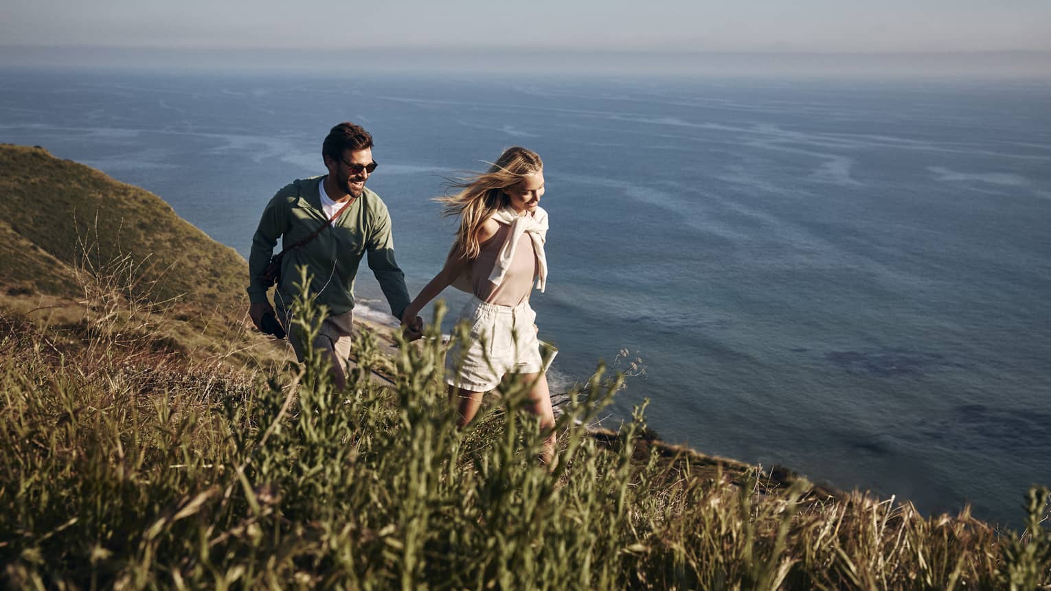 The sun shining on a smiling couple holding hands as they hike along a grassy cliff. Open water ripples below them.