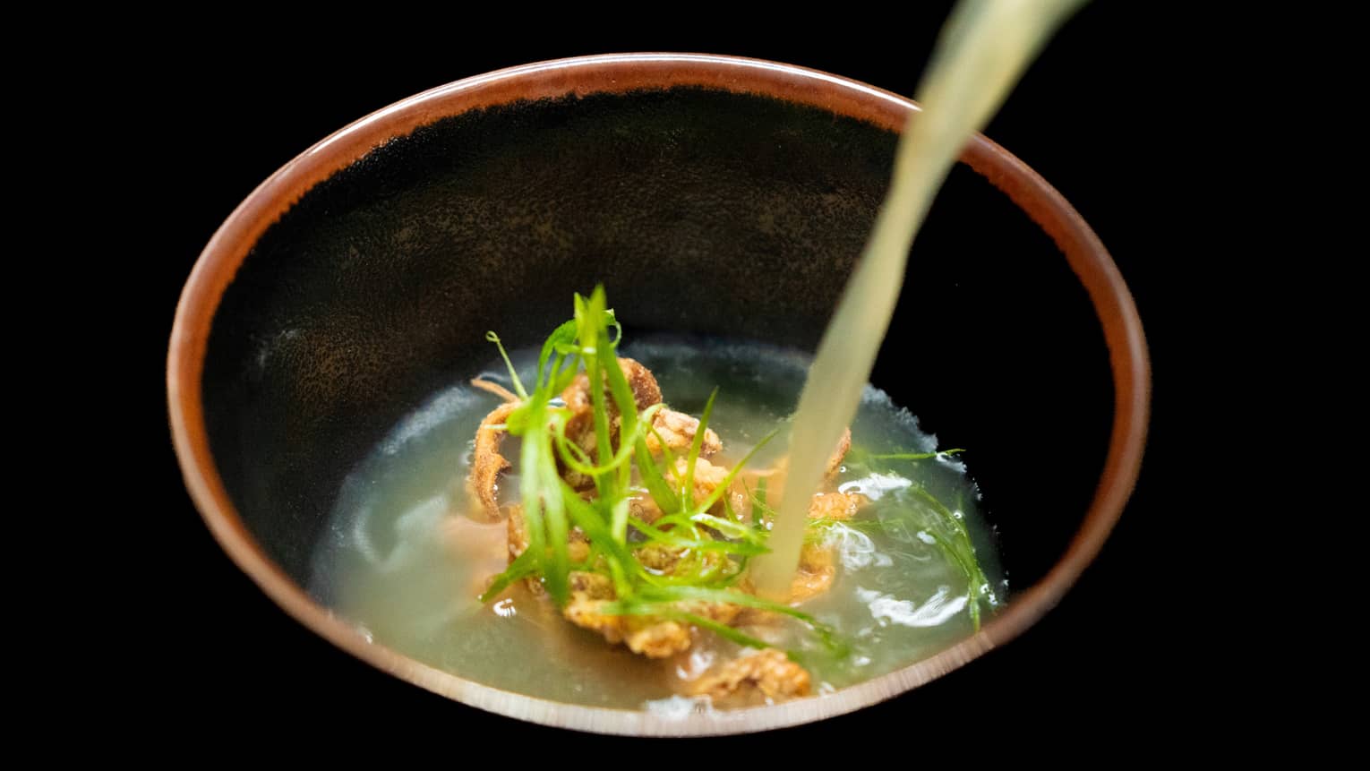 A translucent light-coloured broth is being poured into a deep bowl that also holds food items and garnishes.