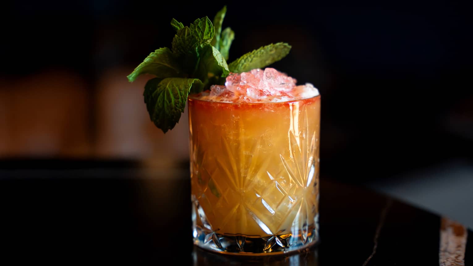 A clear tumbler holds an orangish-pink drink garnished with a sprig of green leaves.