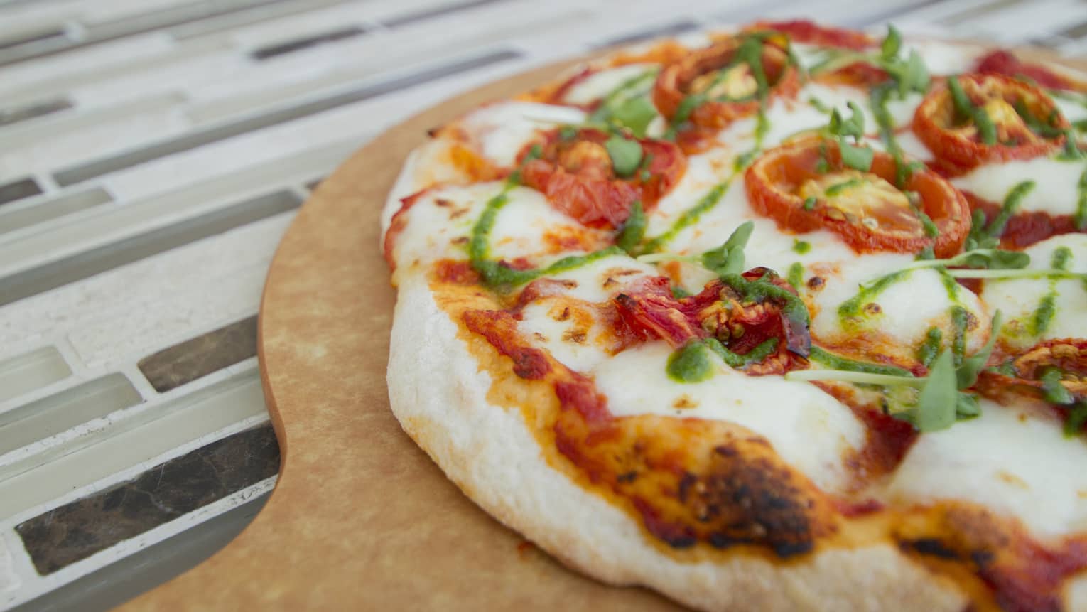A wood-fired marguerita pizza with roasted tomatoes and mozzarella, drizzled with pesto sauce, served on a wood tray. 