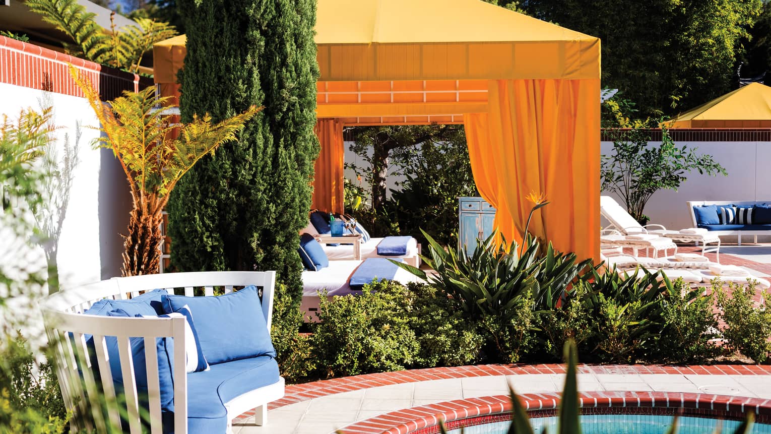 Yellow cabanas, trees on red brick patio by plush blue bench, outdoor swimming pool