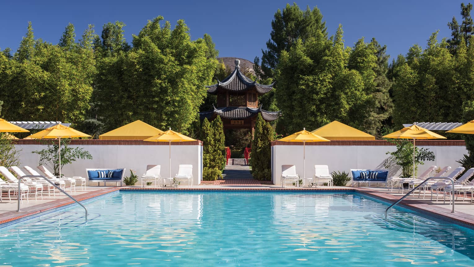 Outdoor swimming pool lined with lounge chairs, yellow umbrellas, path to temple, trees