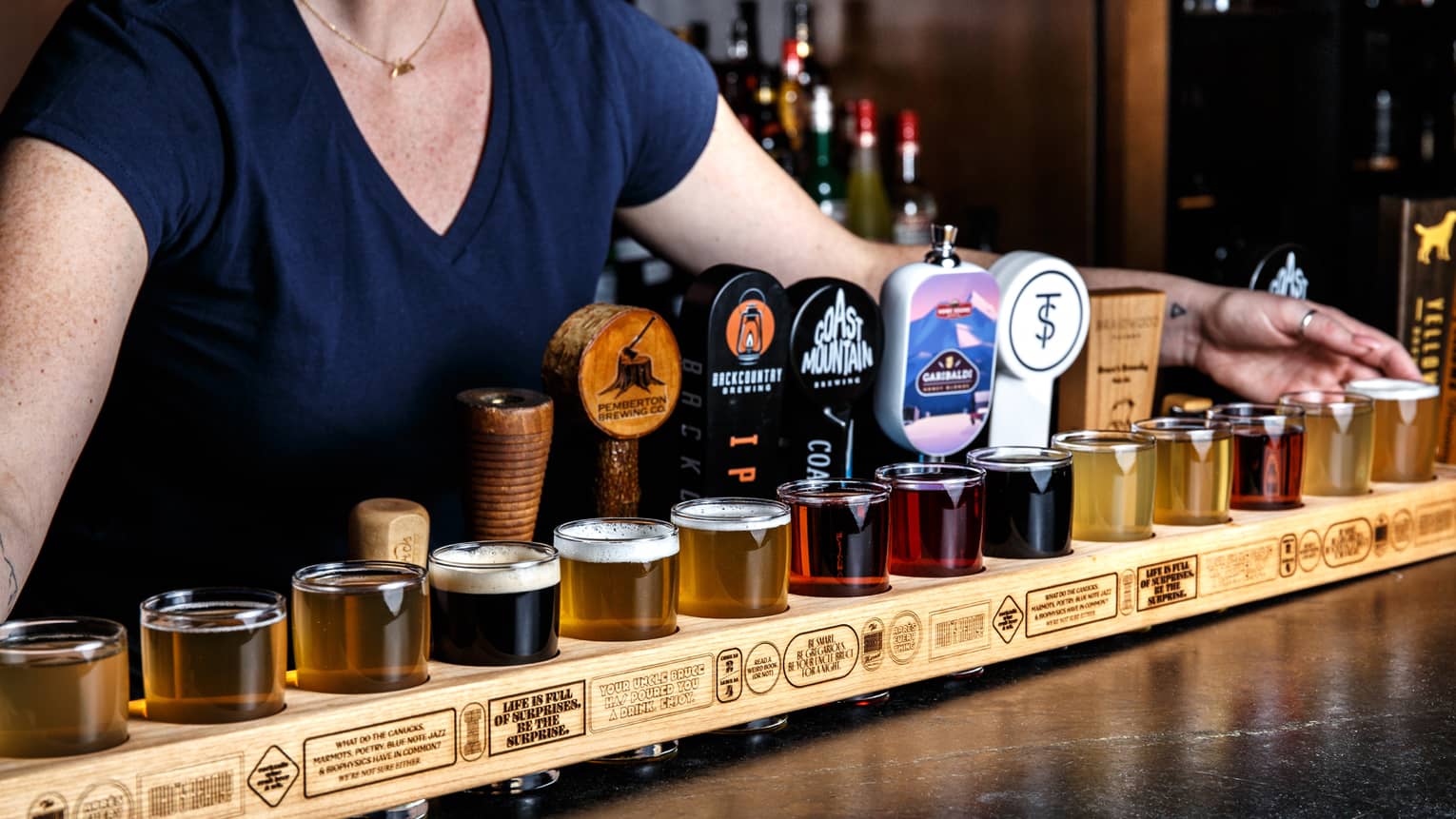 A series of glasses sit on a black table, with a female bartender standing behind.