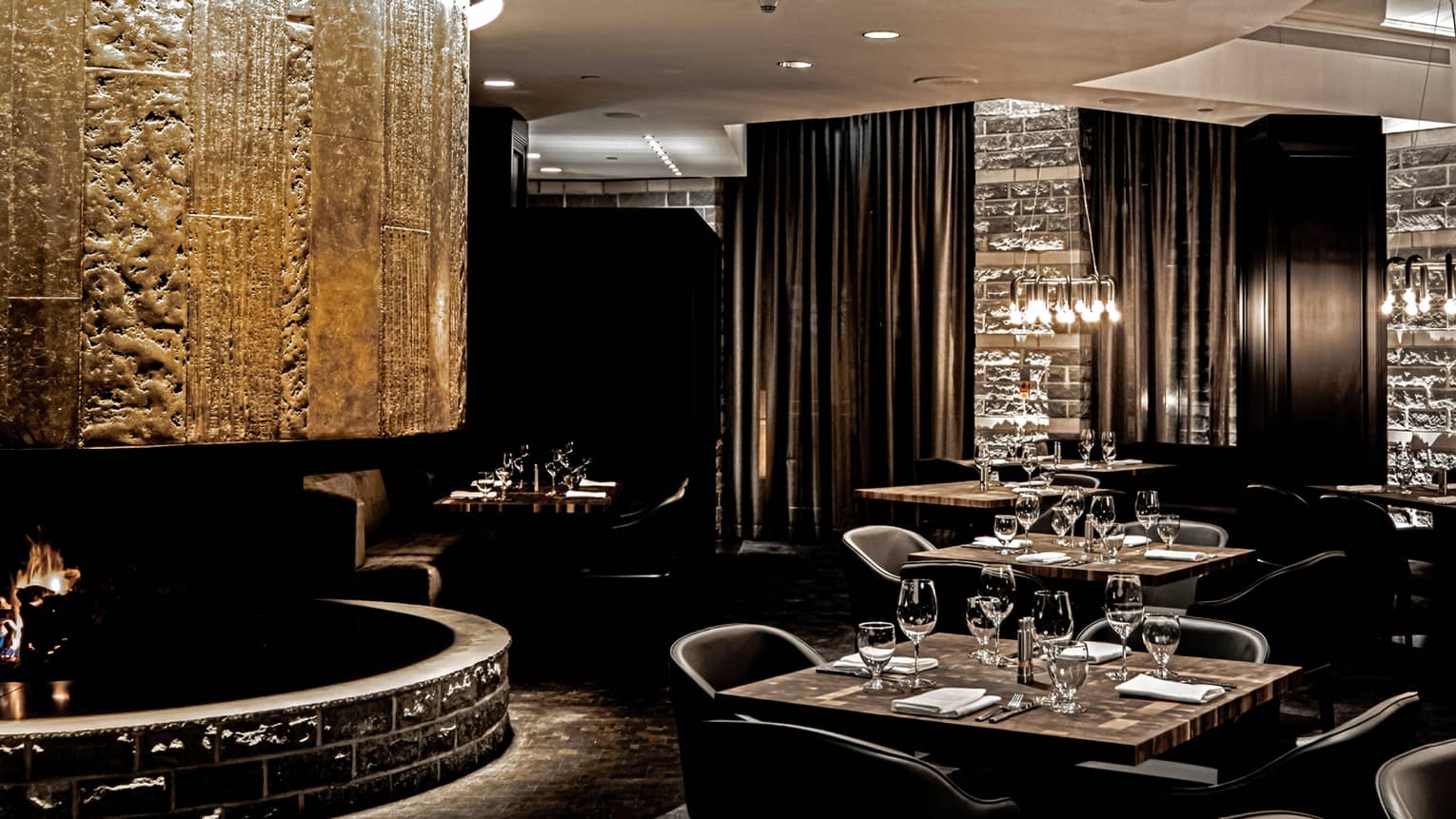 Newly renovated steakhouse with dark wood tables set for four, black curtains, lowlights, fireplace 
