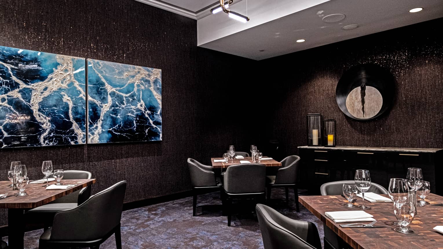 An upscale restaurant with dark walls and abstract art, three tables each set for four with leather club chairs.