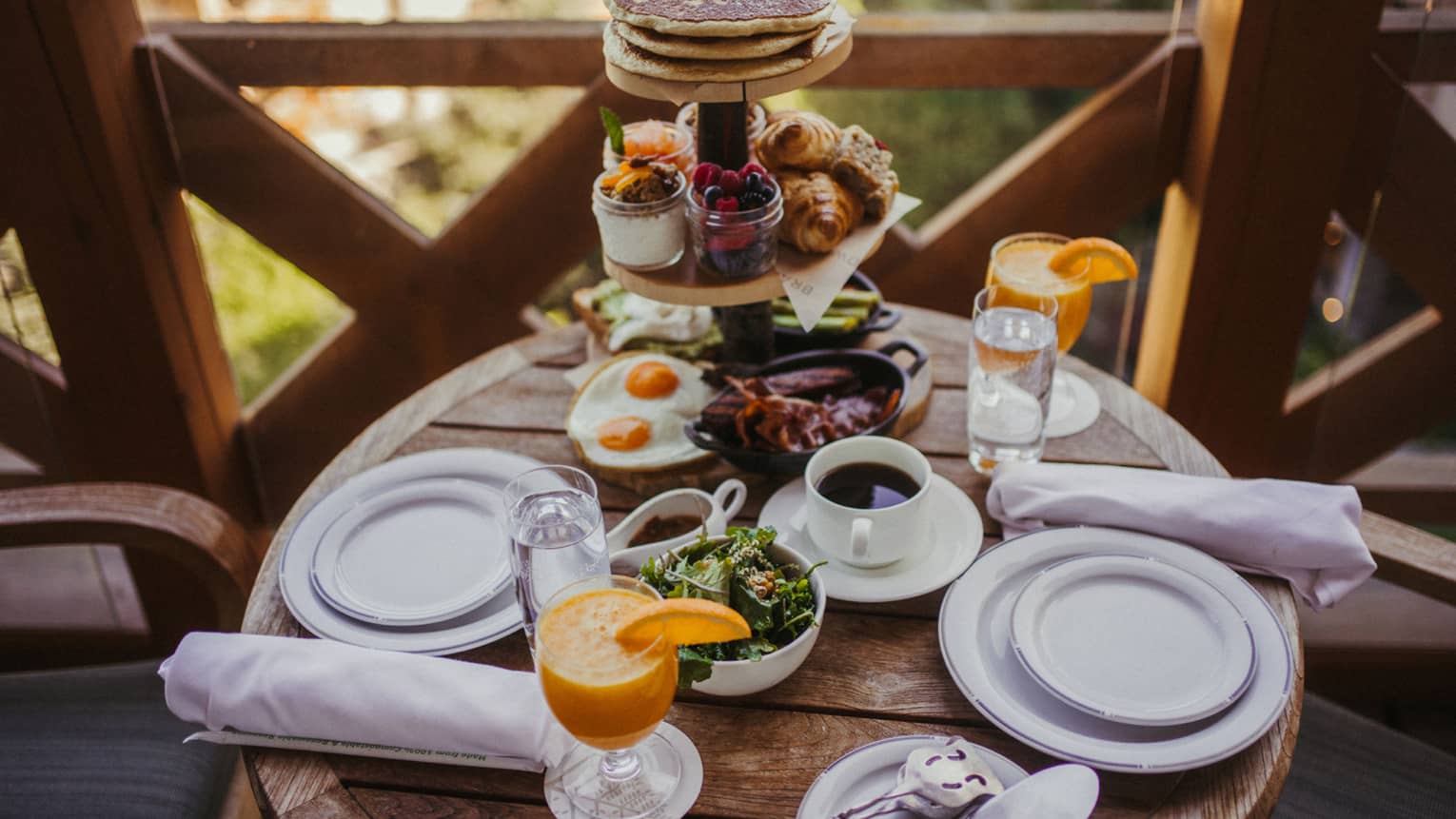 A full breakfast spread of orange juice, coffee, pancakes and eggs on white porcelain plates, served on a wooden balcony.
