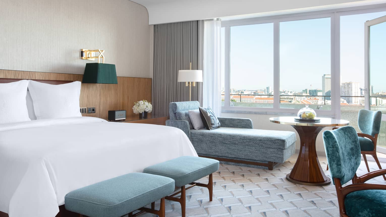 Guest room with white bed, blue benches at foot, blue chaise longue, tall windows with view