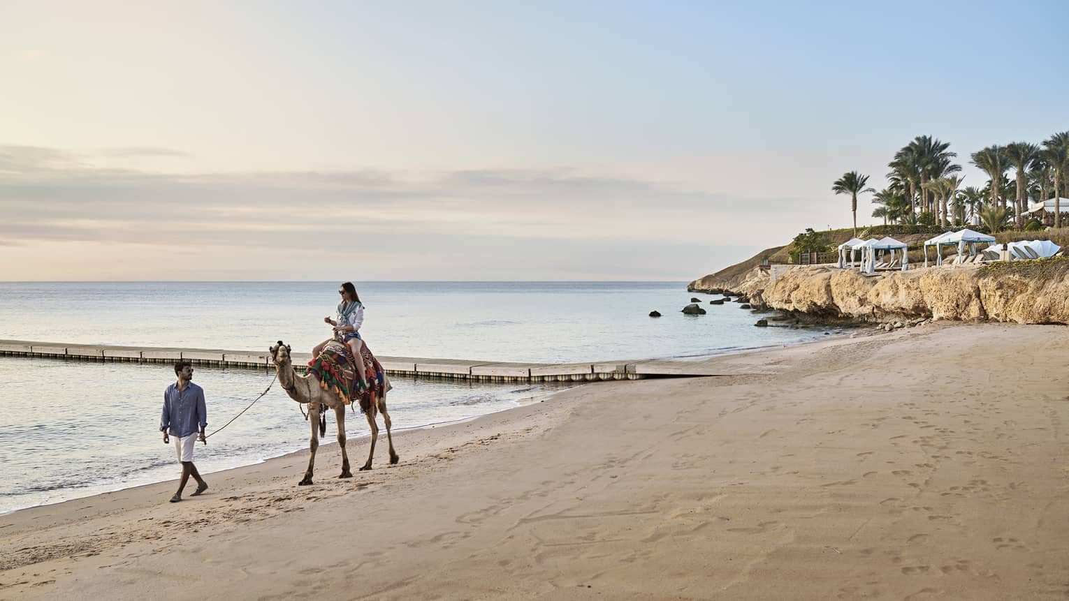 A woman rides a camel, led by a man, along the beach at Four Seasons Sharm El Sheikh