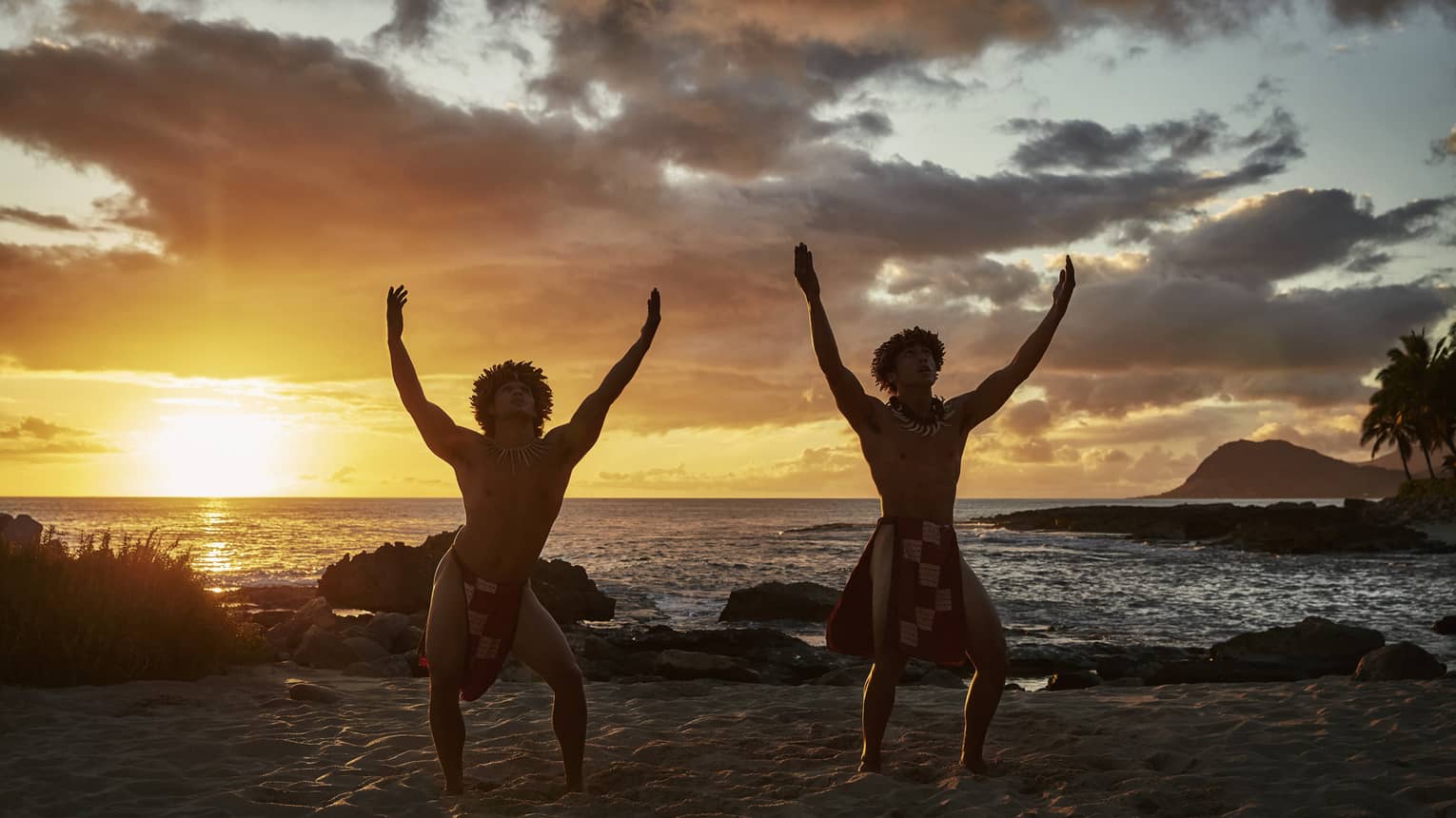 Two hula dancers on the beach, sinewy arms raised above their heads, in silhouette against dramatic fiery clouds at sunset.