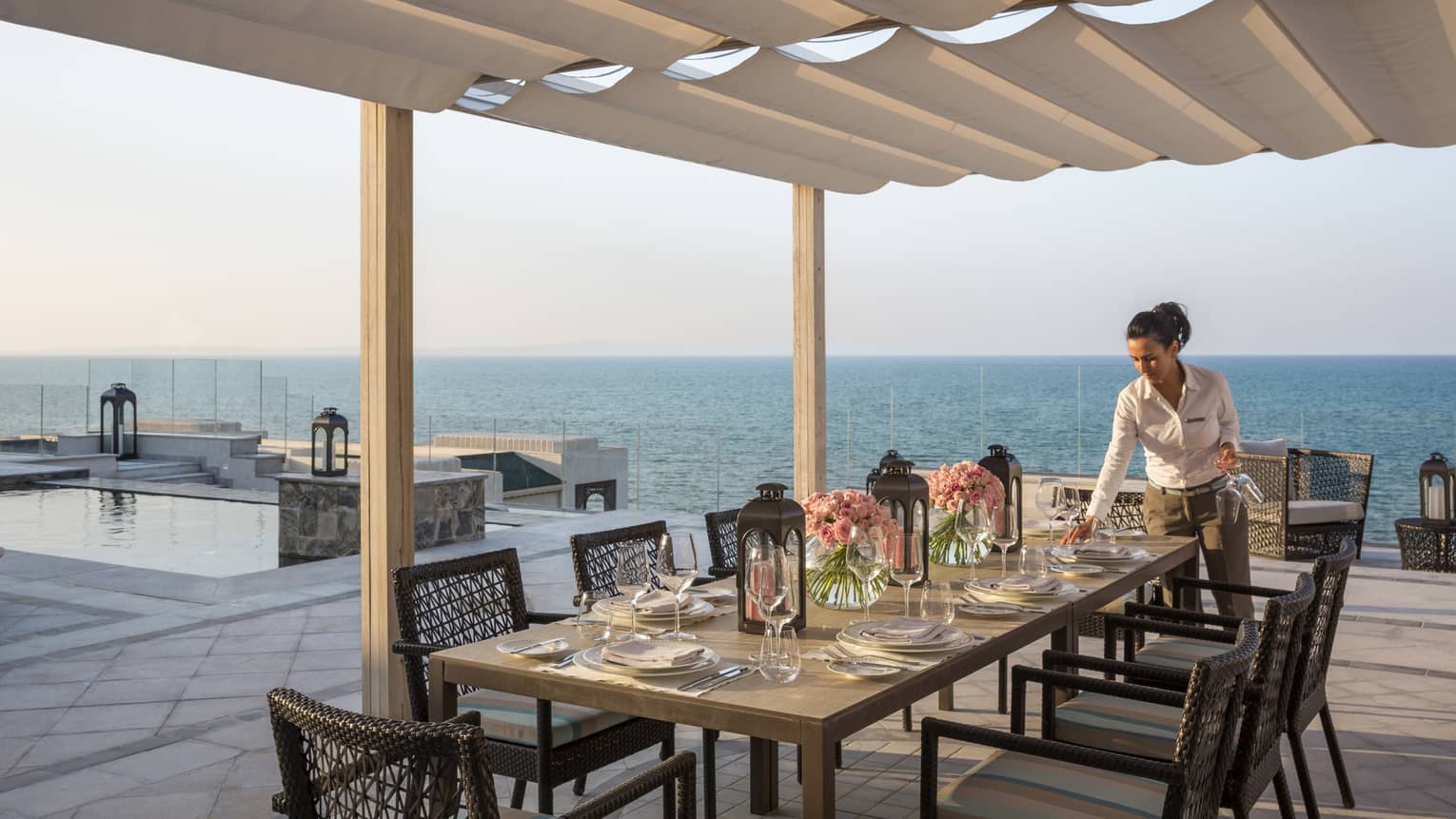Woman sets table with flowers and lanterns on private rooftop patio overlooking pool and ocean views