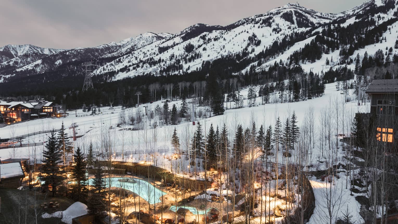 Aerial view of illuminated blue outdoor swimming pool through trees on snowy hill at dusk