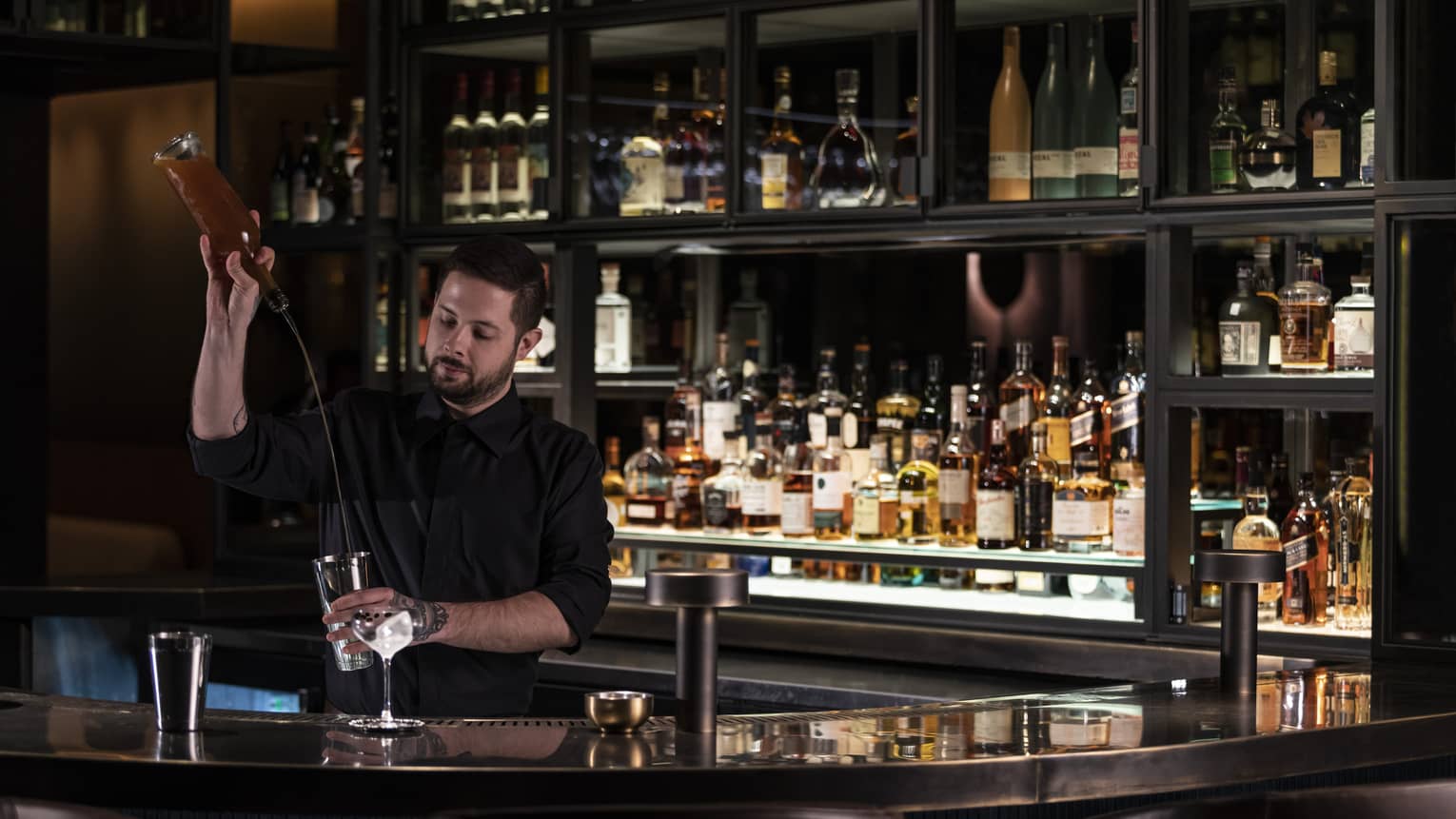 Mixologist pours alcohol from bottle into cocktail shaker behind sleek bar 